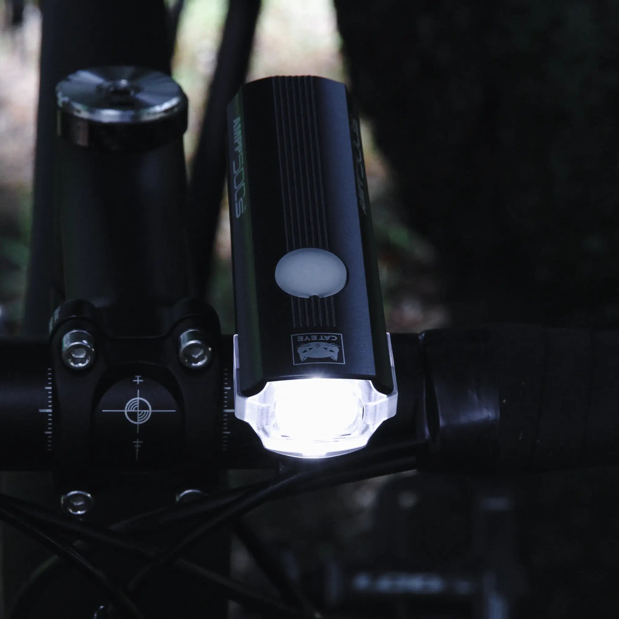 A close-up of a bicycle handlebar shows the CATEYE AMPP500S headlight with OptiCube™ lens, mounted near the stem using a FlexTight™ bracket, shining brightly forward in a low-light outdoor setting.