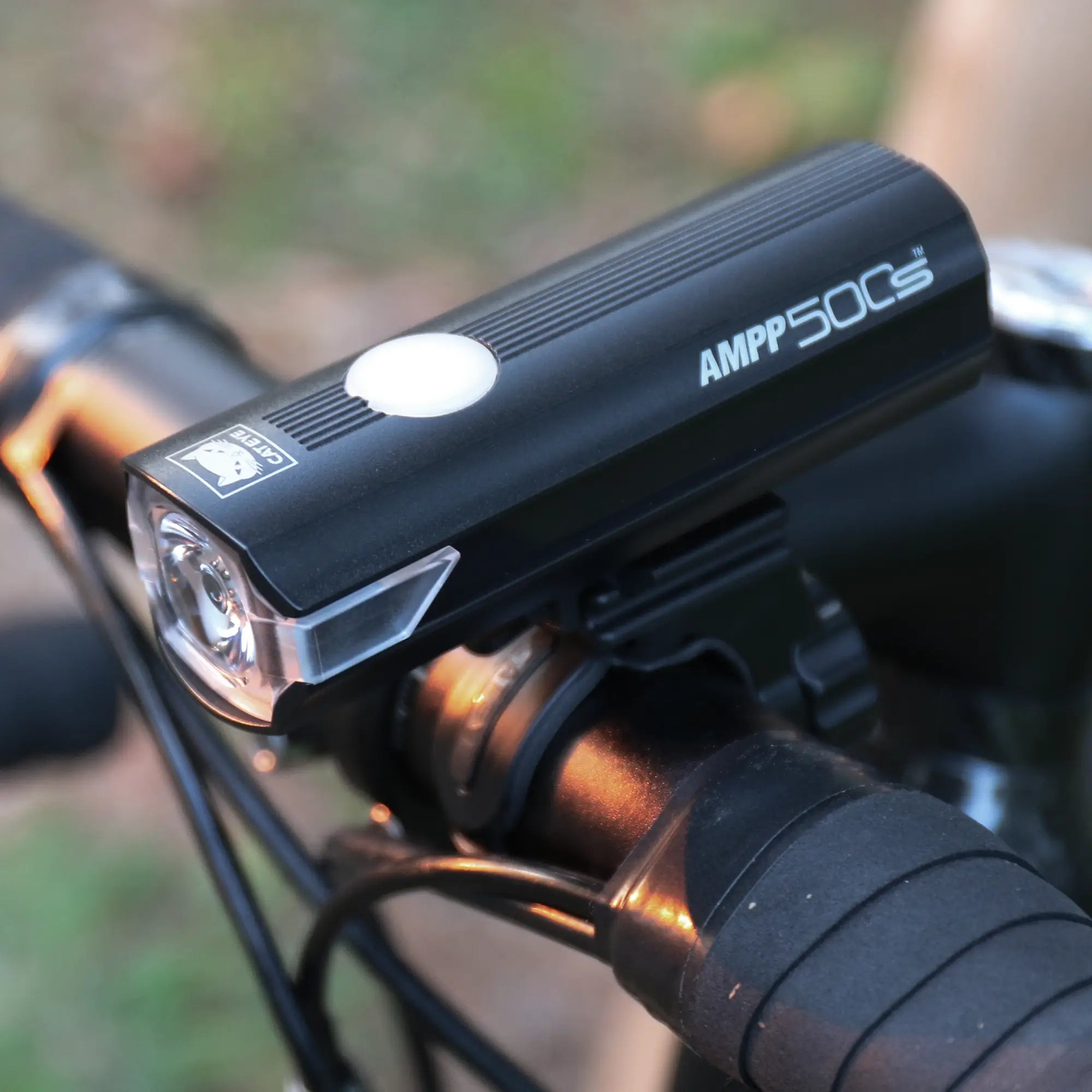 A close-up of a CATEYE AMPP500S black bicycle headlight with an OptiCube™ lens, mounted on a handlebar with a FlexTight™ bracket, set against a blurred outdoor background.