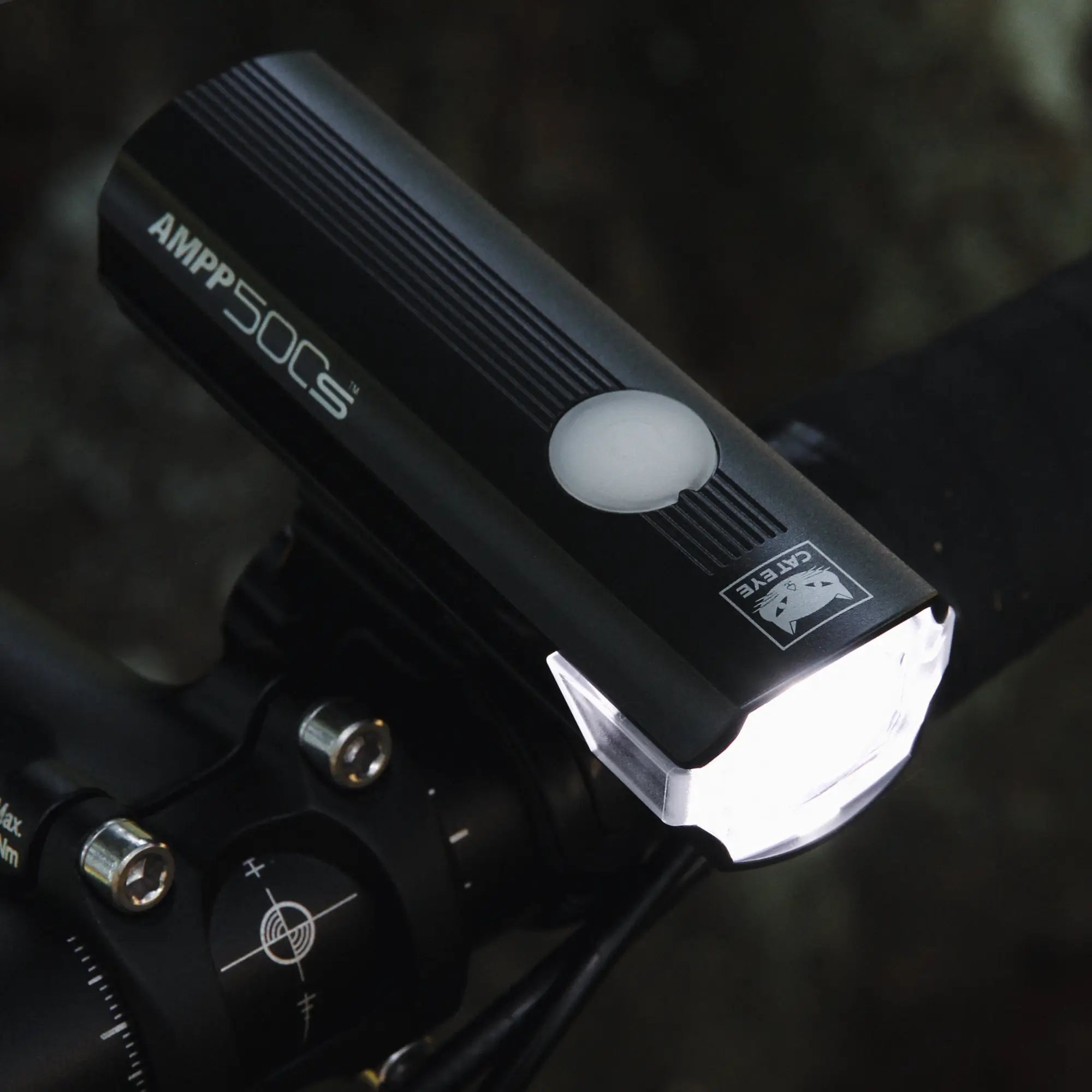 Close-up of the CATEYE AMPP500S bicycle headlight with OptiCube™ lens, mounted via FlexTight™ bracket on handlebars, shining bright white light in a dimly lit setting.