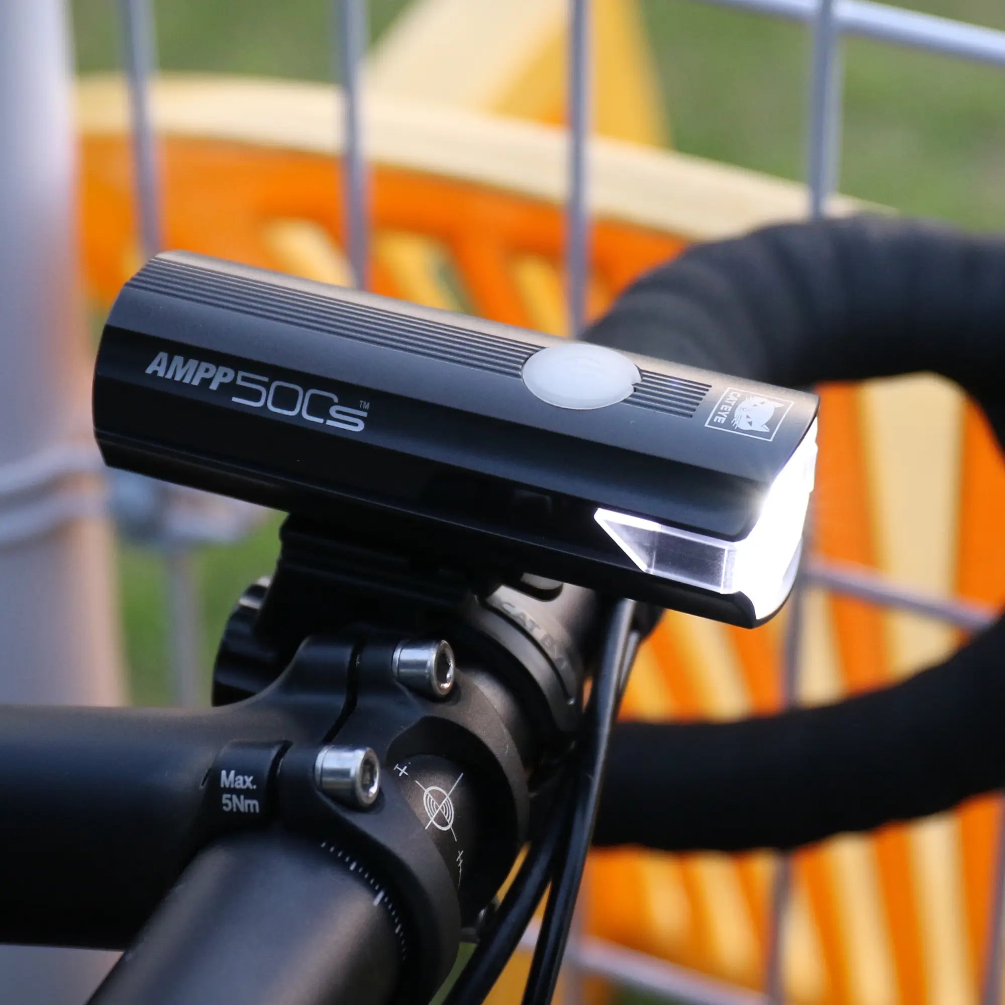 A close-up of a black CATEYE AMPP500S bicycle headlight with its white OptiCube™ lens illuminated, mounted on handlebars using a FlexTight™ bracket. A metal fence and orange structure appear blurred in the background.