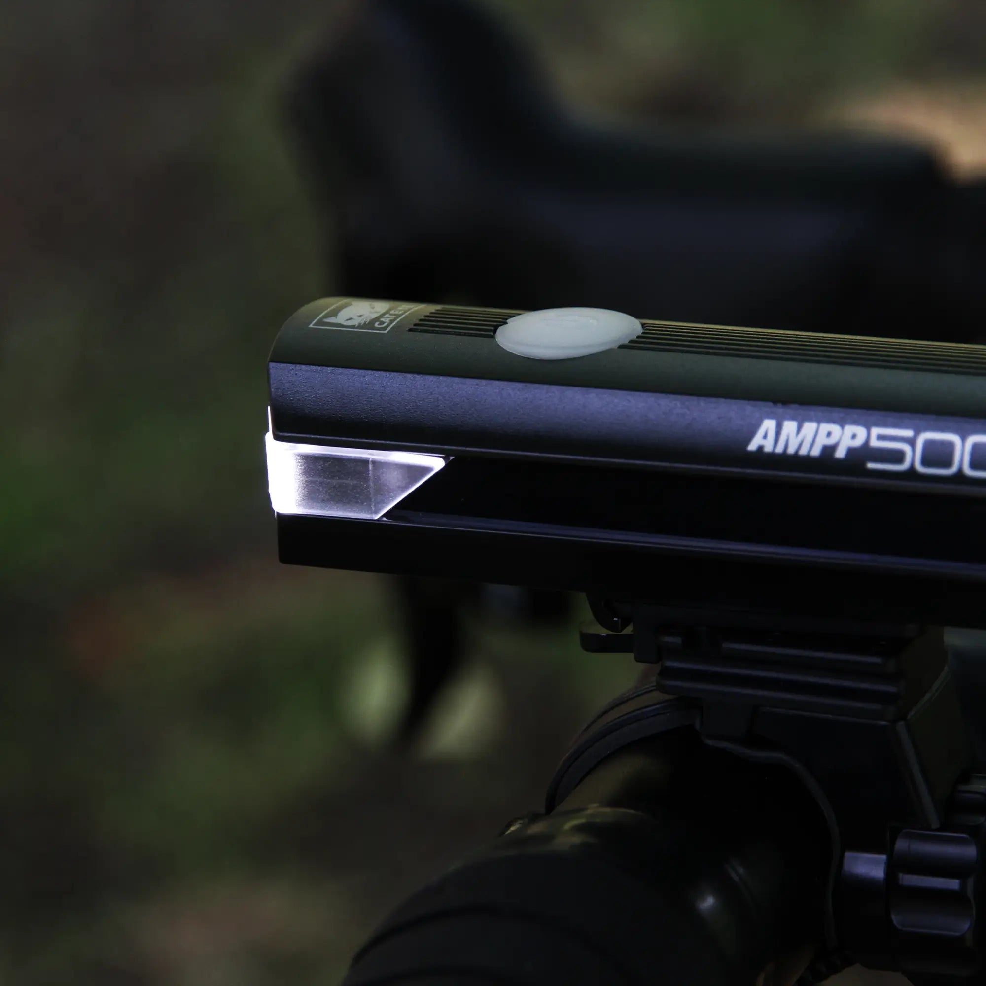 Close-up of a CATEYE AMPP500S black bike headlight with OptiCube™ lens, mounted on handlebars via FlexTight™ bracket, glowing white against a blurred outdoor backdrop.