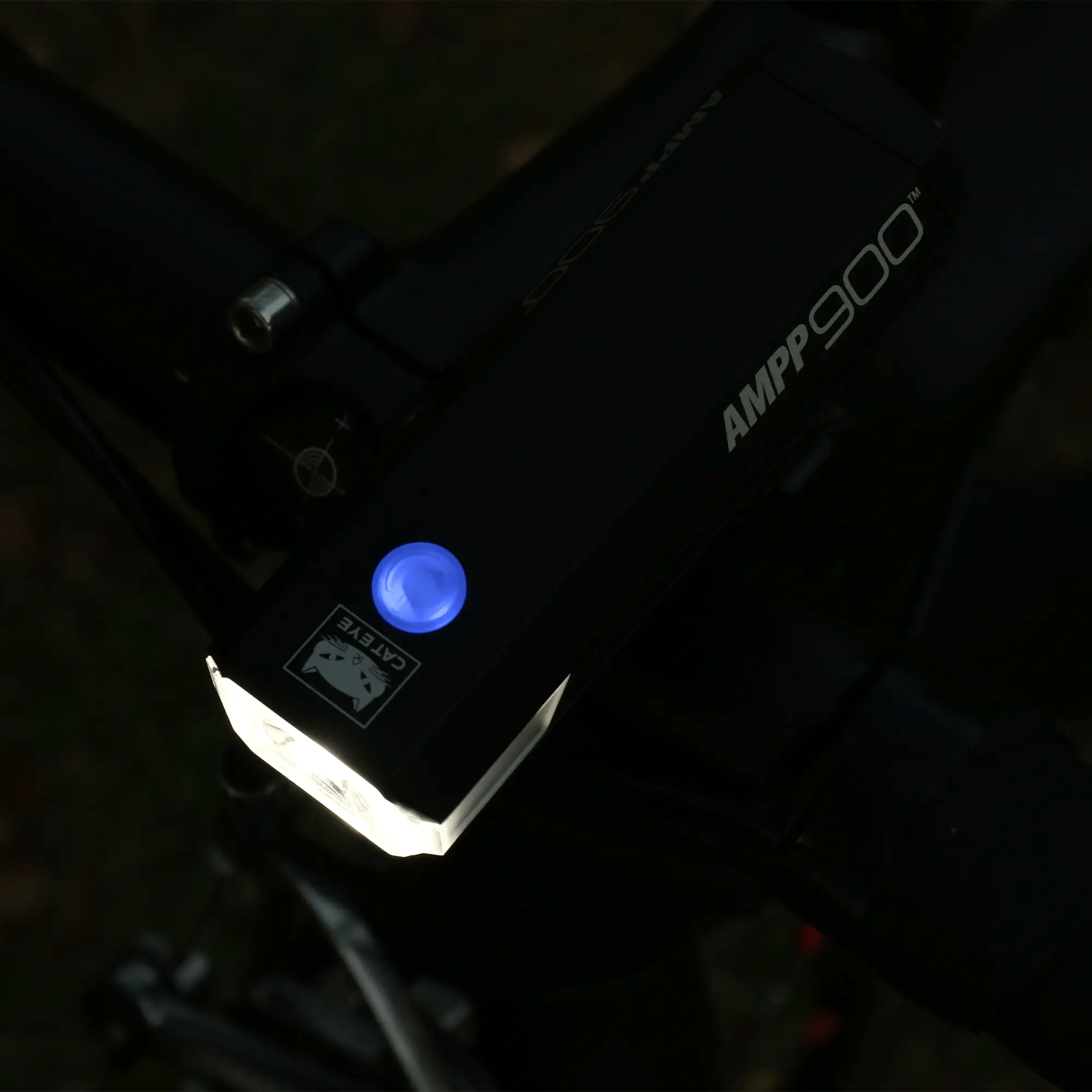 A close-up of the CATEYE AMPP900 bike headlight mounted on handlebars showcases its OptiCube™ lens, illuminated blue power button, and 900-lumen beam shining in a dim setting.