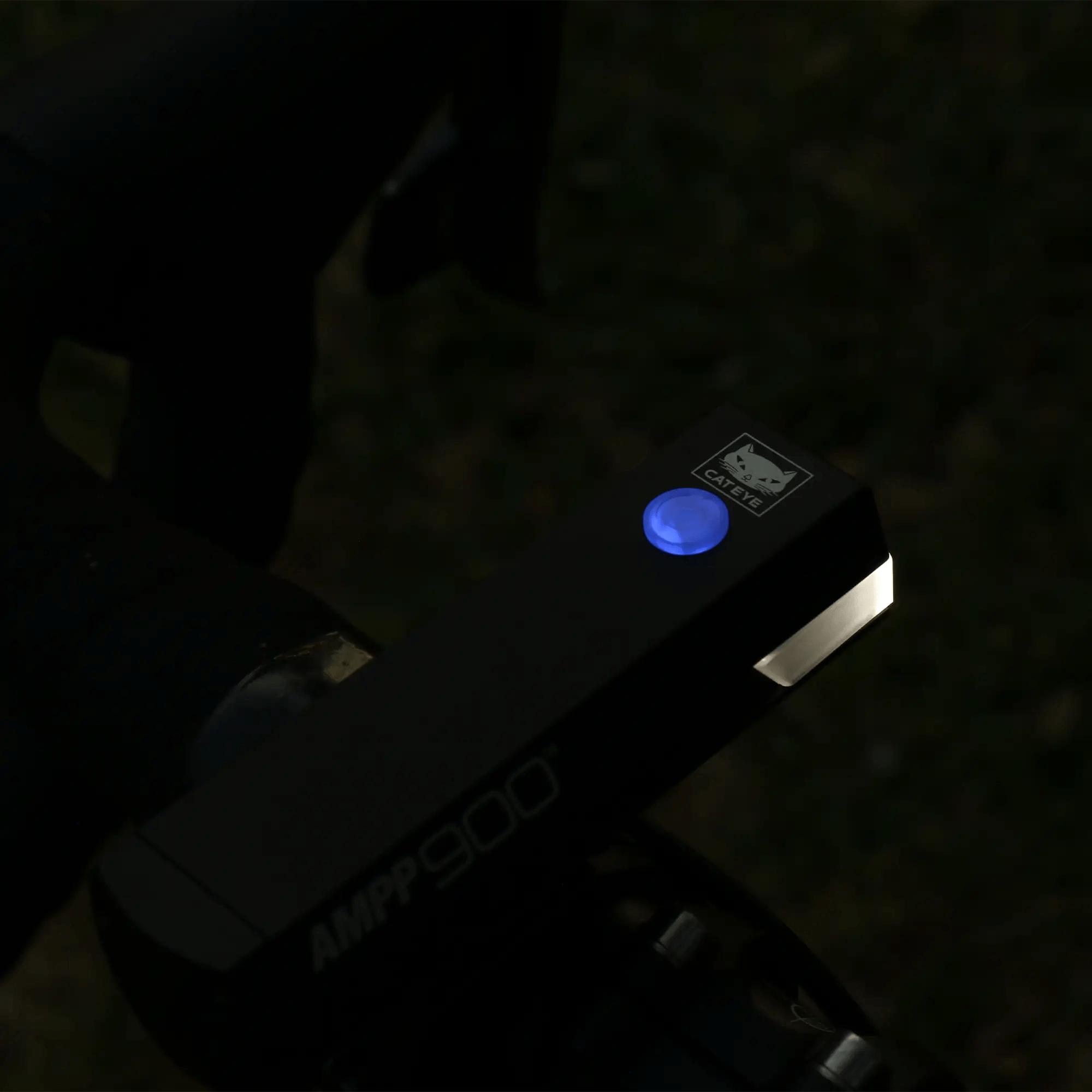 A close-up of a bicycle handlebar showcases the CATEYE AMPP900 bike light with OptiCube™ lens technology, its 900 lumen beam and glowing blue power button highlighting the CATEYE logo against a dark, blurred background.