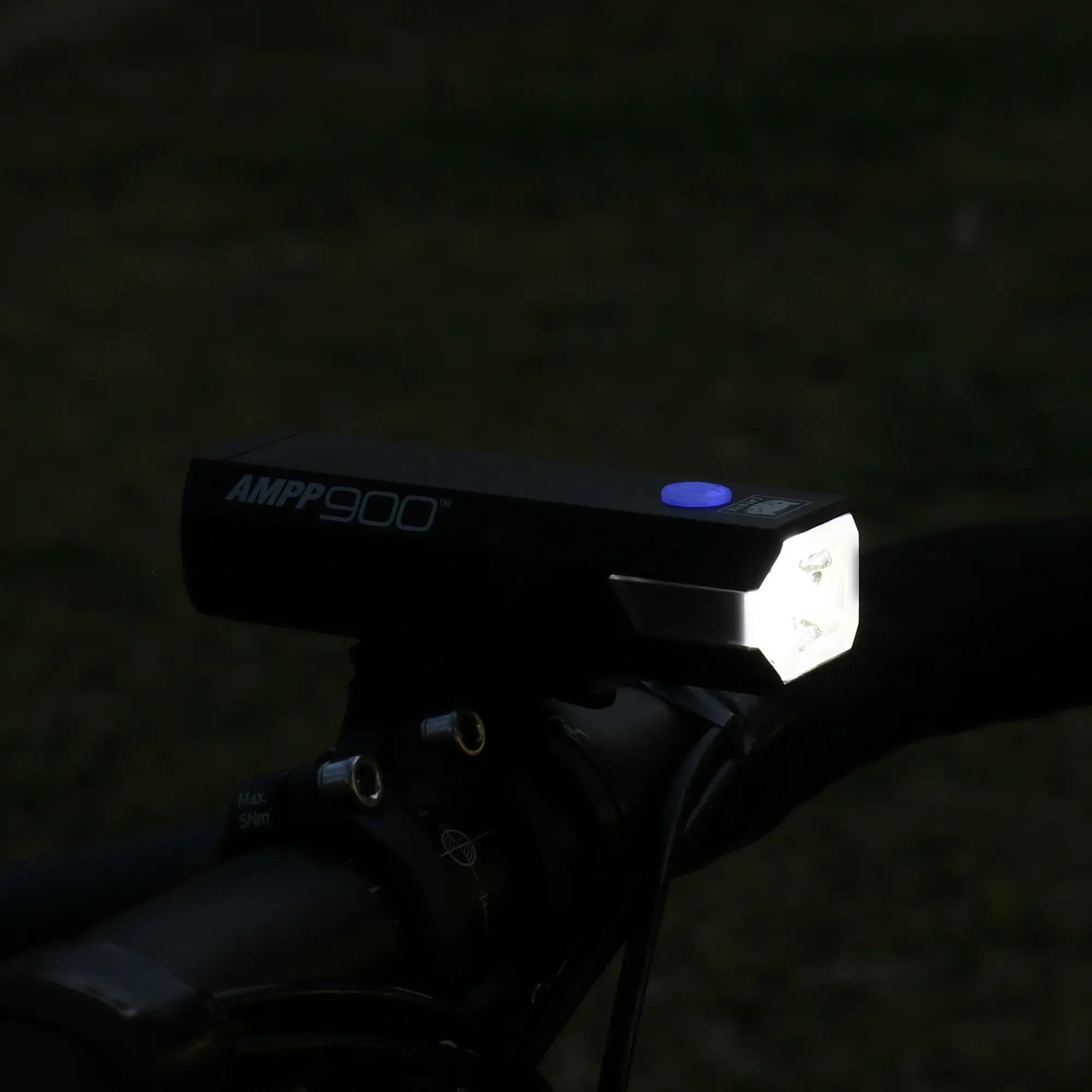 A black CATEYE AMPP900 bike headlight with a glowing white front and blue button is mounted on a handlebar, shining a powerful 900 lumen beam to brighten the dim outdoor scene.