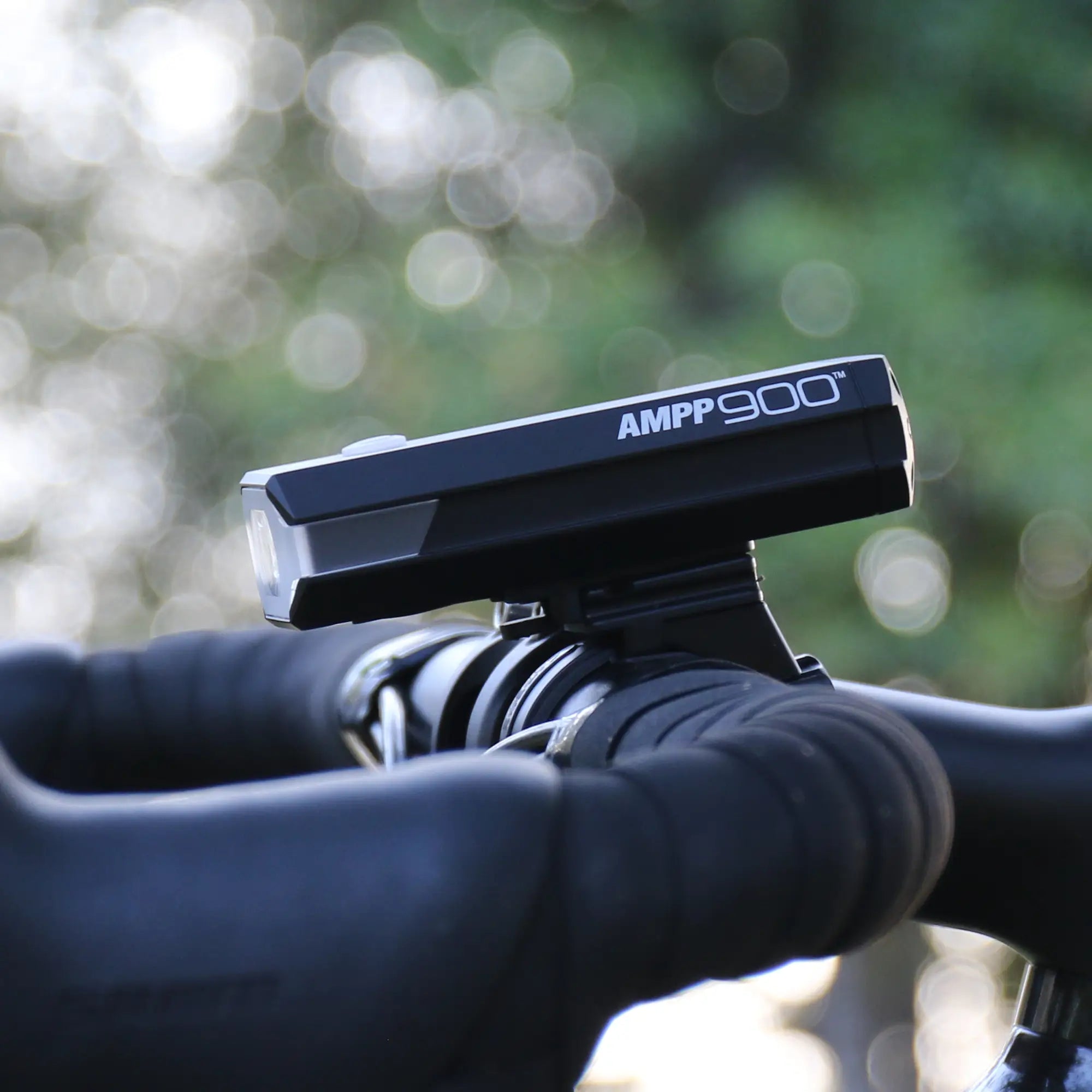 Close-up of the CATEYE AMPP900 black bicycle headlight featuring OptiCube™ lens technology, mounted on handlebars with a blurred green and white outdoor background.