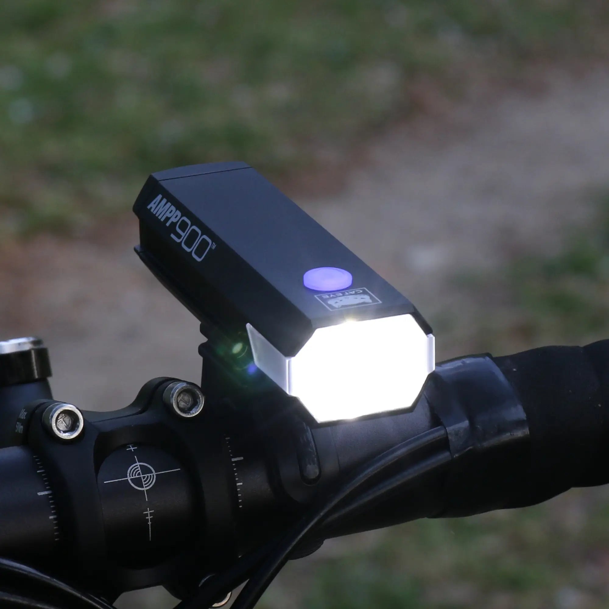 A CATEYE AMPP900 bike headlight with OptiCube™ lens technology is mounted on the handlebars, casting a powerful 900-lumen beam. The background features an out-of-focus path and lush greenery.