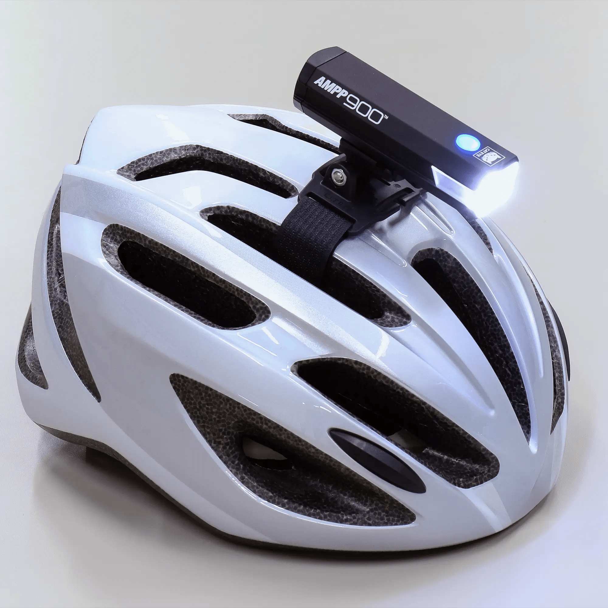 A white bike helmet with black vents is equipped with a mounted black CATEYE AMPP900 front light, featuring OptiCube™ lens technology, a blue glowing button, and a bright 900-lumen white beam.
