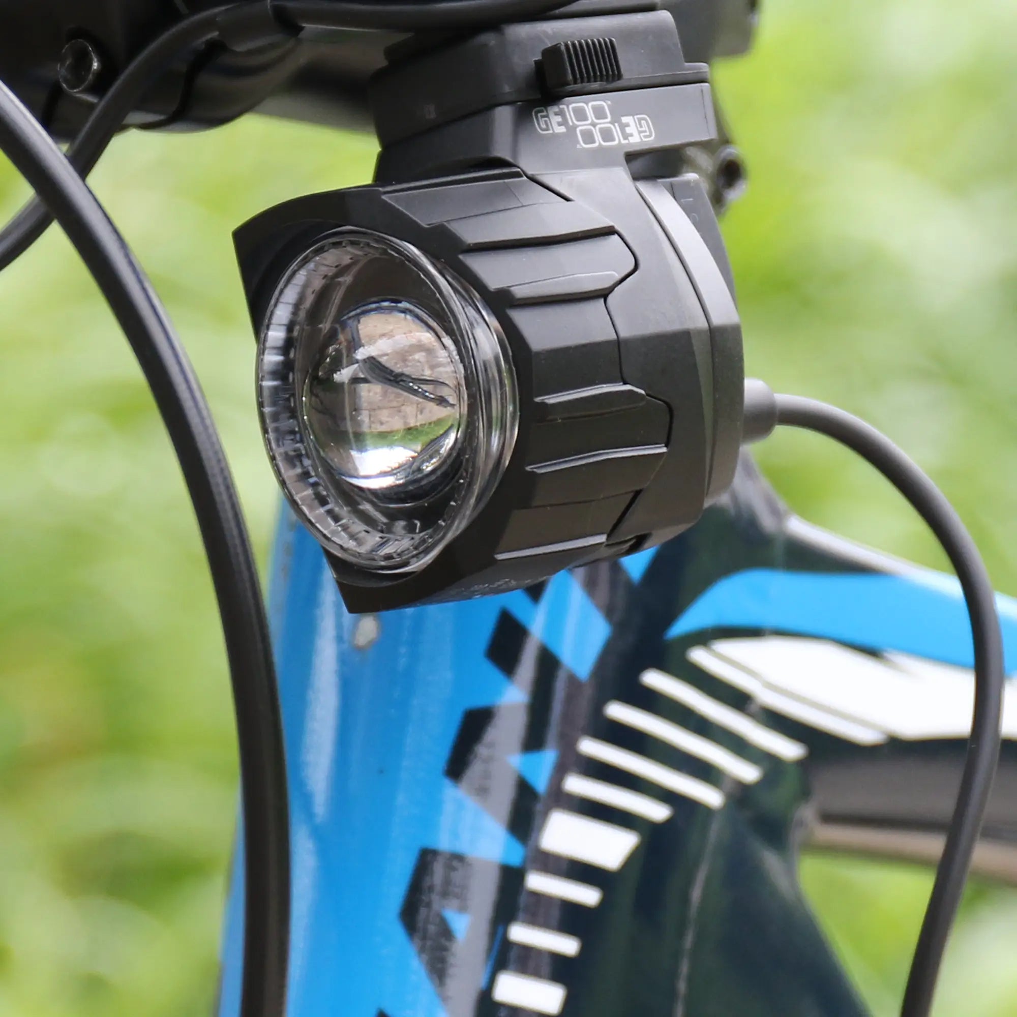 Close-up of a CATEYE GE100 Connect E-Bike Headlight (StVZO certified, 100 Lux) mounted on the front frame of a blue and white bike, visible cables, with a blurred green background.