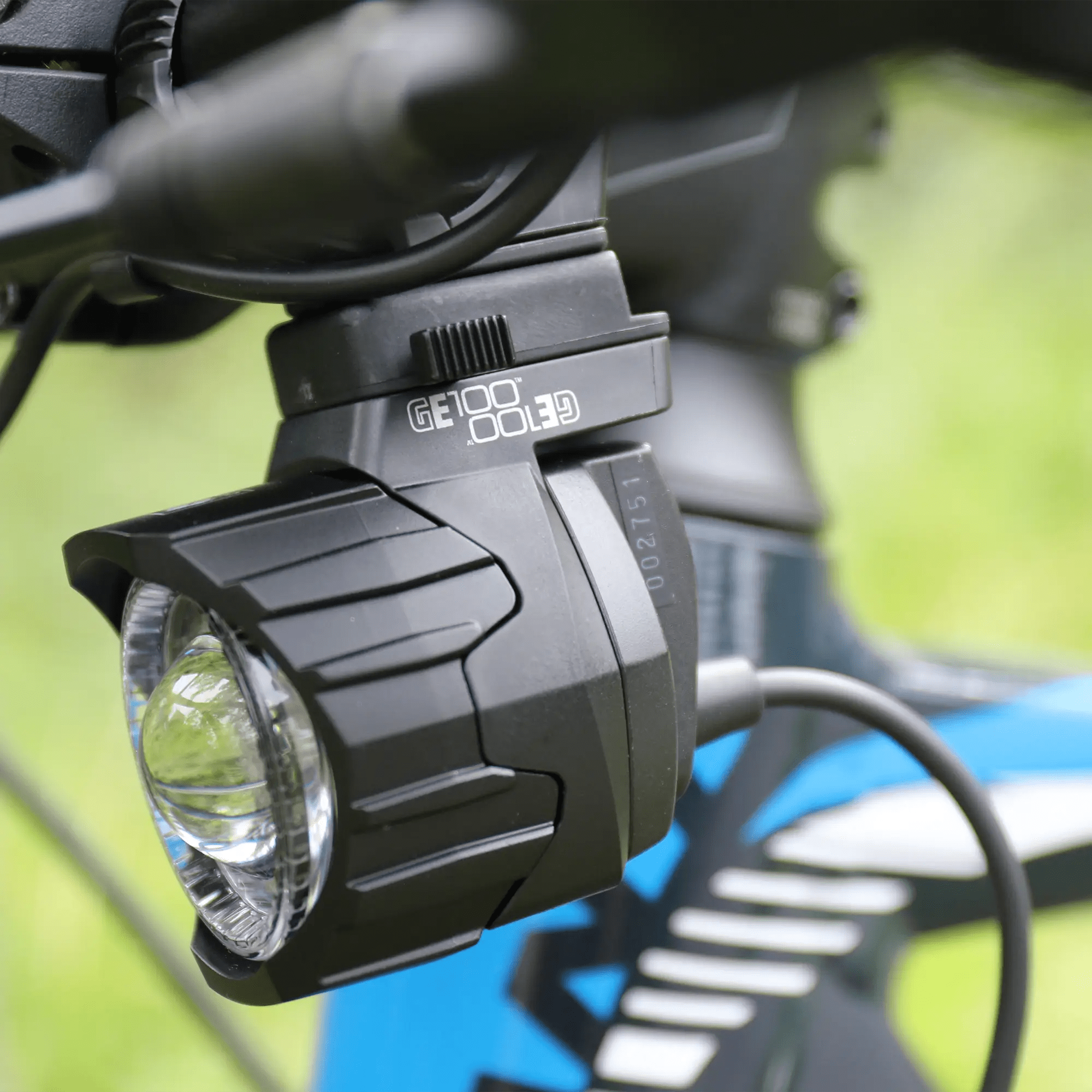 A close-up of a CATEYE GE100 Connect E-Bike Headlight, 100 Lux and StVZO certified, mounted on the handlebars of a blue bike, with a blurred green background.