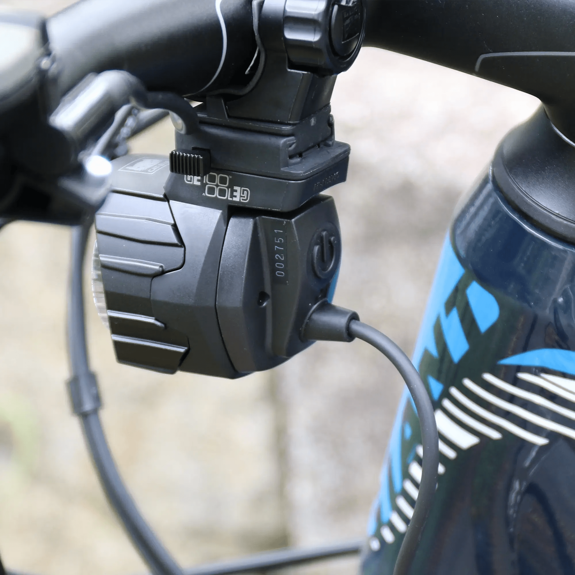Close-up of a CATEYE GE100 Connect E-Bike Headlight, StVZO certified and cable-connected, mounted on a bicycle handlebar beside a blue and black bike frame with white graphics.