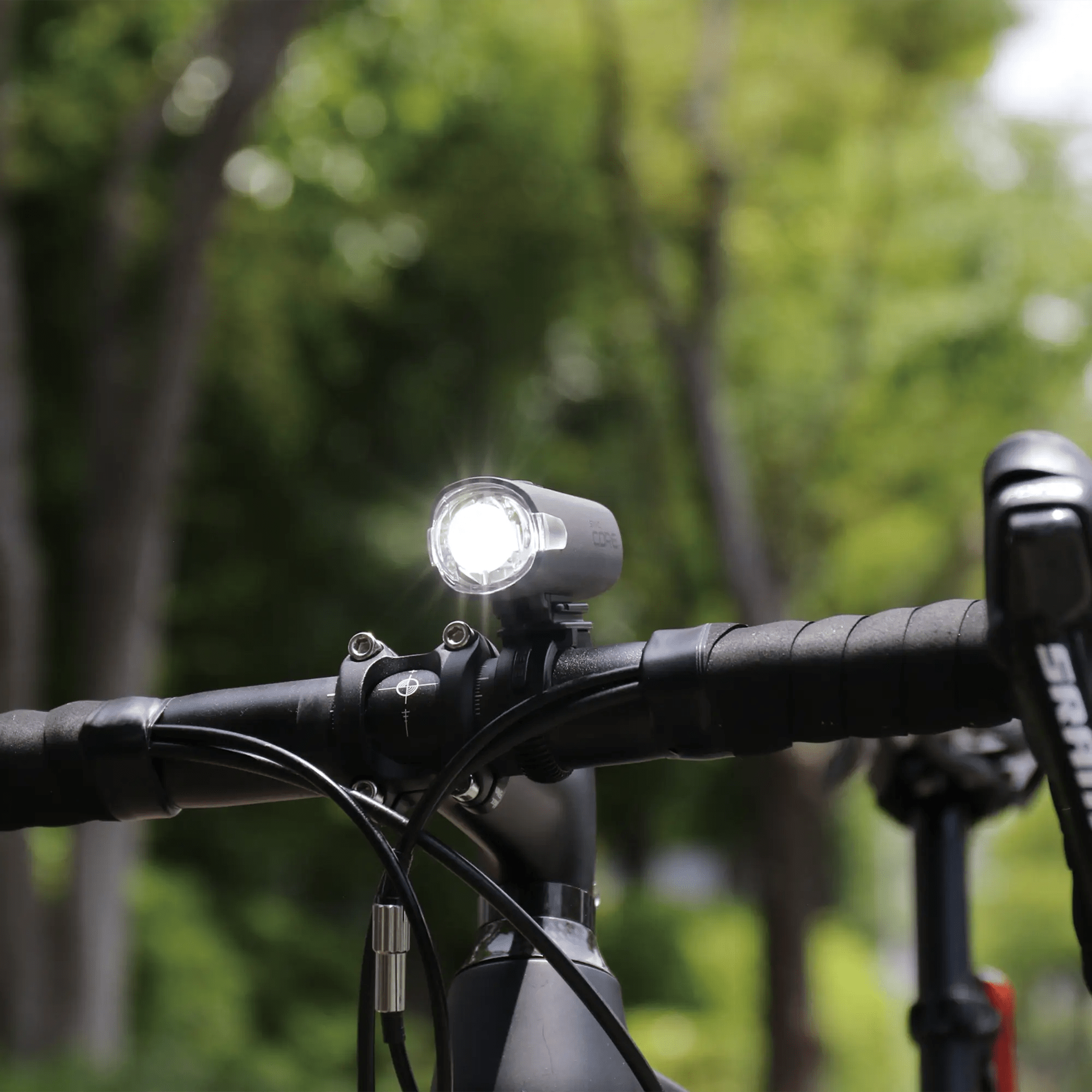 A CATEYE SYNC Core front light turned on is mounted on a bicycle handlebar, providing 360-degree visibility, with green trees blurred in the outdoor background.