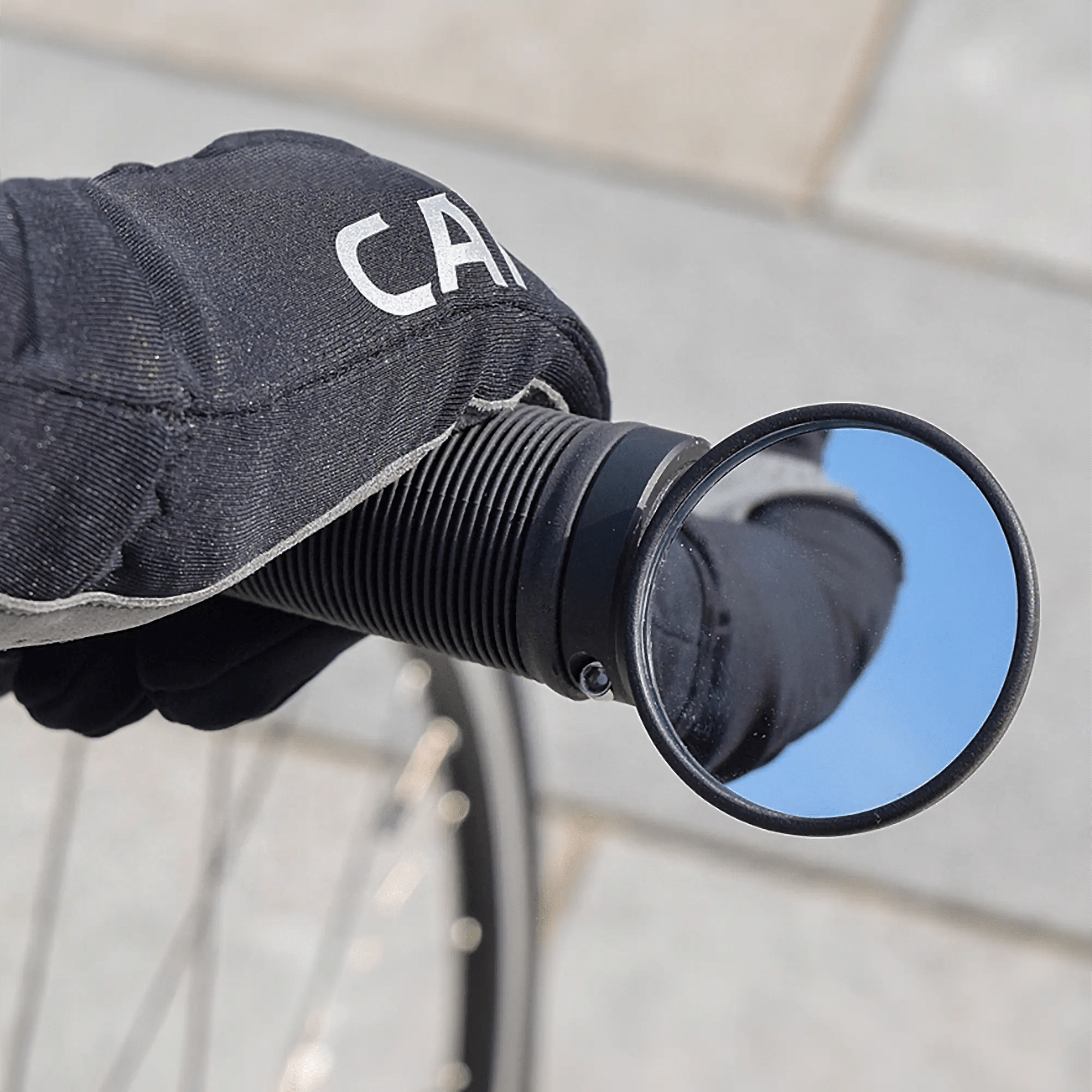 A gloved hand grips a bicycle handlebar equipped with the CATEYE BM-45 Bar End Mirror, which reflects the blue sky above large, light-colored paving stones.