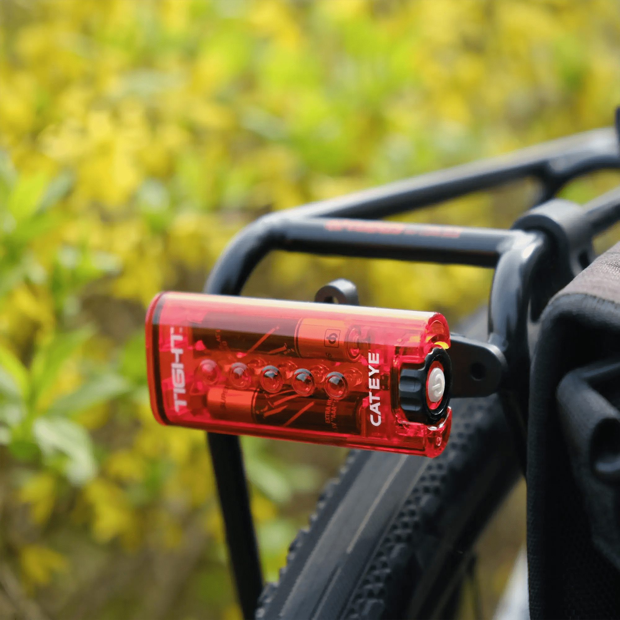 A close-up of a red CATEYE Snap System rear light mounted on a black Rear Rack Bracket, with green and yellow blurred foliage in the background.