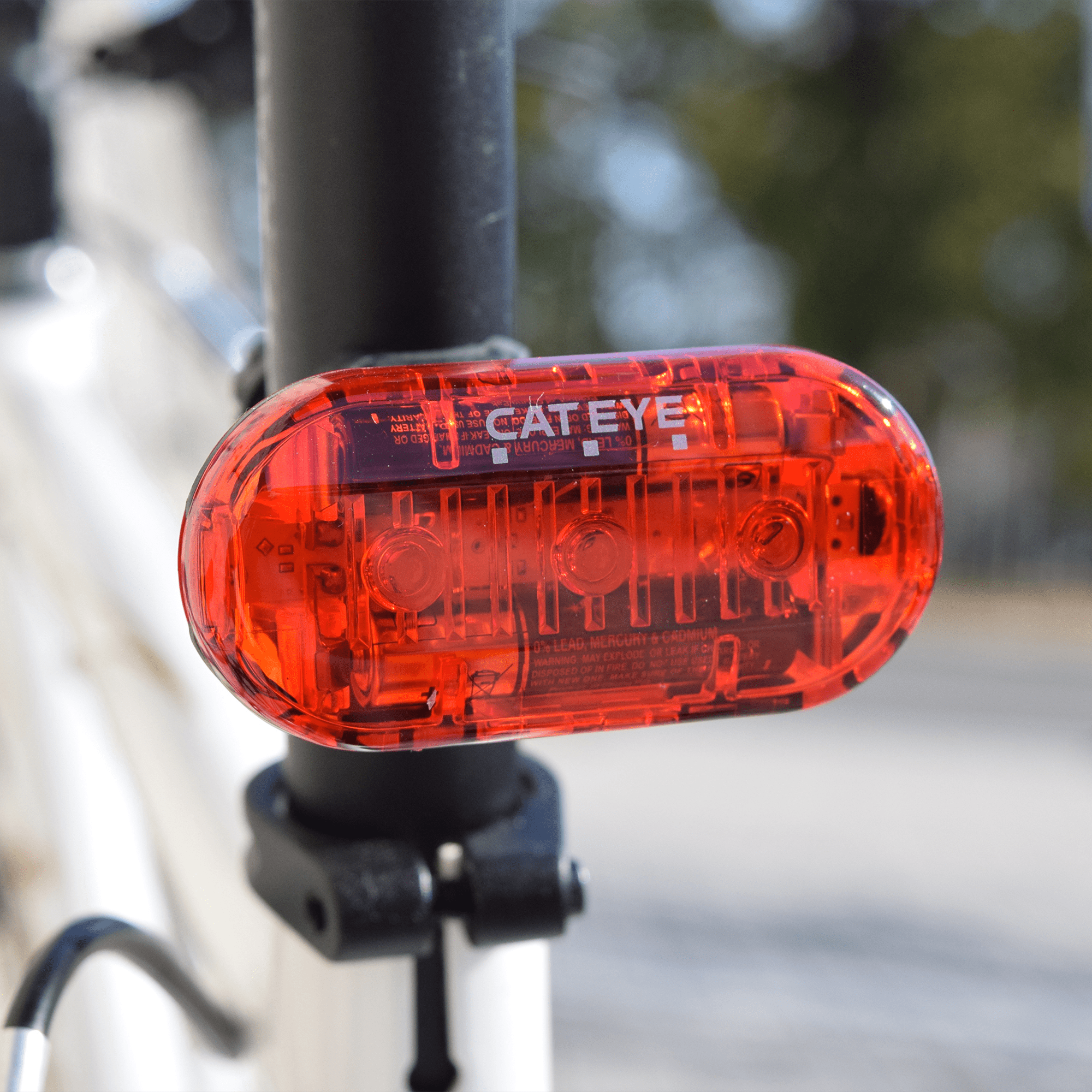 Close-up of a CATEYE OMNI 3 cycling safety light with OptiCube™ lens, mounted on a seat post. Its transparent housing shows internal parts, while the blurred outdoor background highlights its modern design.