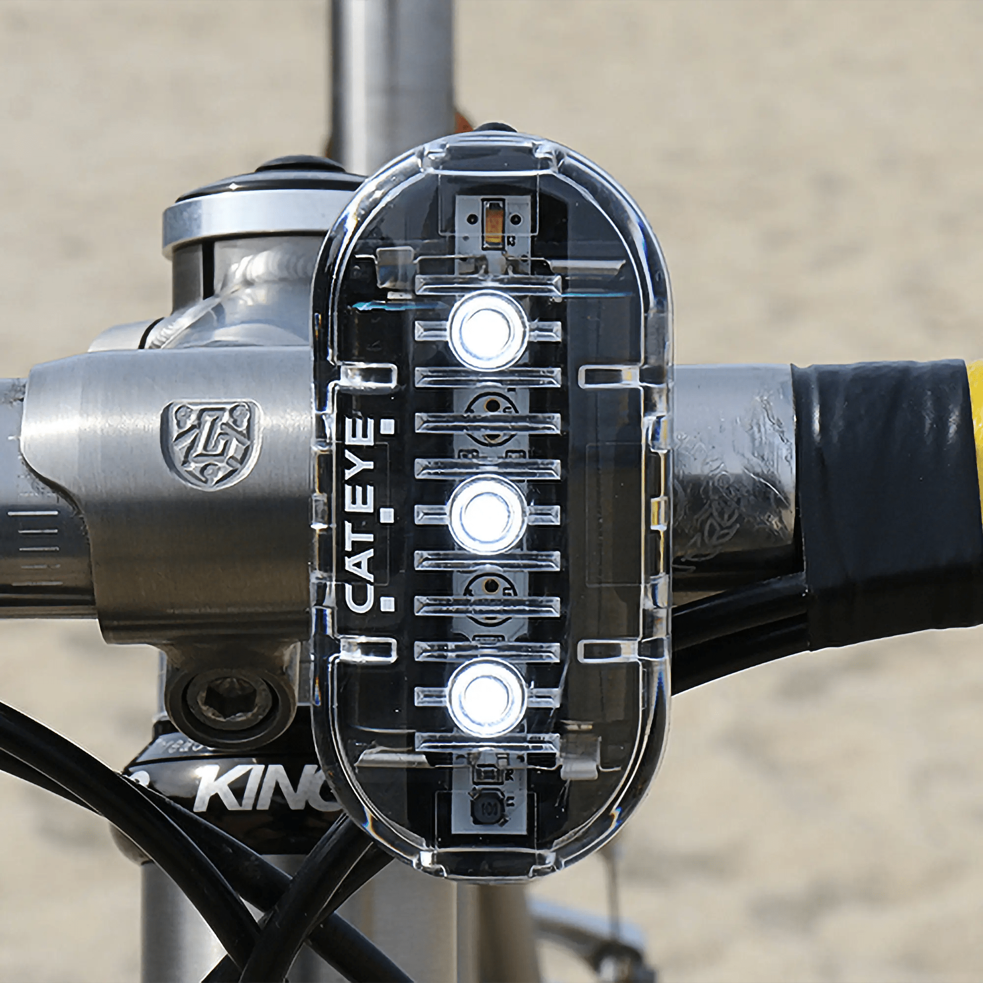 A CATEYE OMNI 3 cycling safety light with four white LEDs and OptiCube™ lens is mounted on bicycle handlebars, with a KING logo visible on the metal stem in the background.