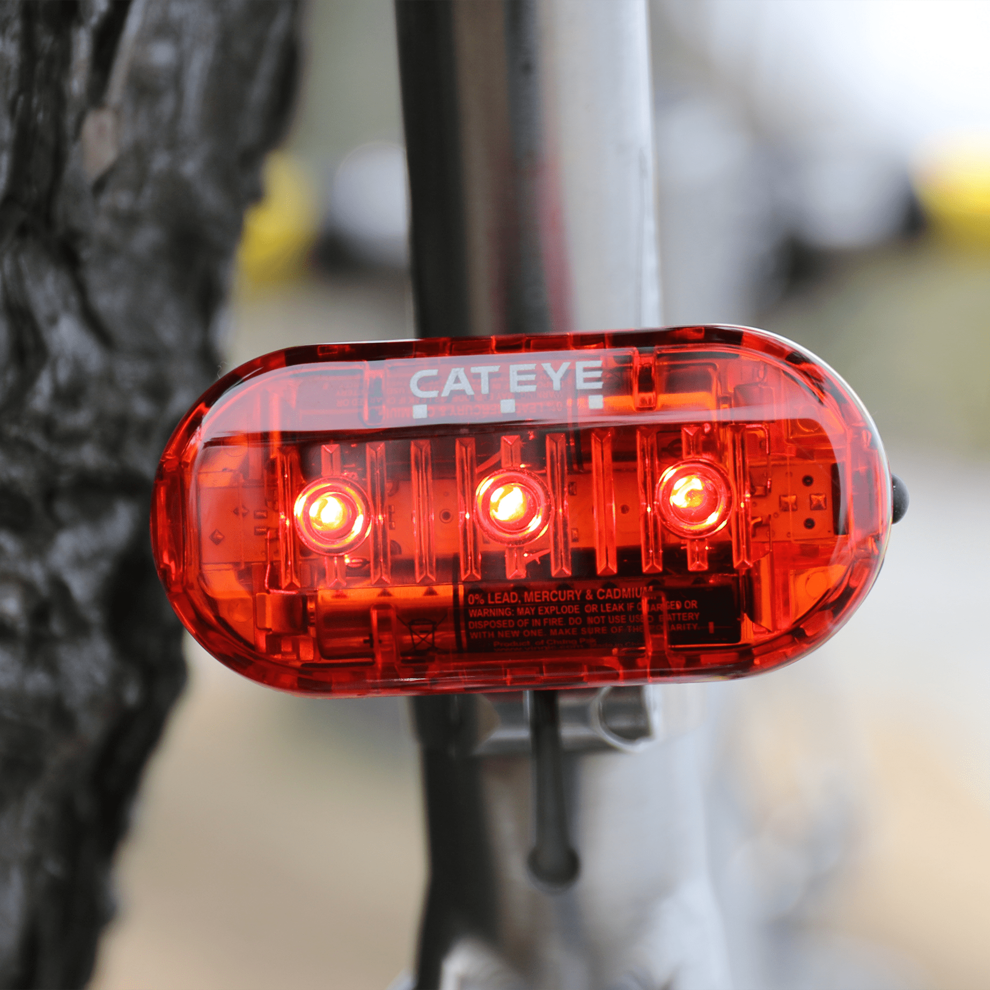 A CATEYE OMNI 3 red cycling safety light with OptiCube™ lens technology is mounted on a silver bike seat post, shining with three LEDs. The background is blurred, showing a tree trunk and indistinct outdoor scenery.