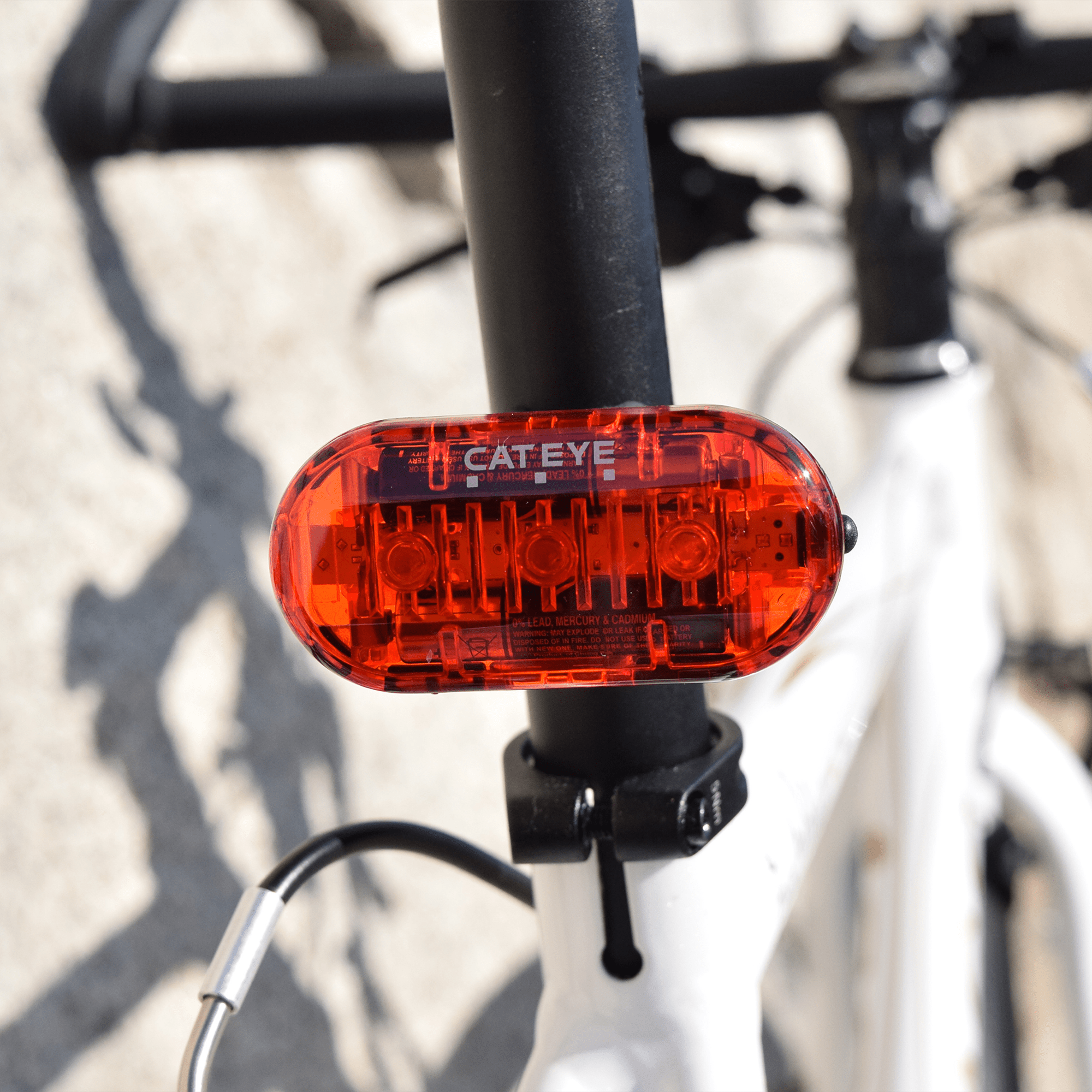 A close-up of the CATEYE OMNI 3 red cycling safety light with OptiCube™ lens, mounted on a white bikes seat post; handlebars and a stone wall are blurred in the background.