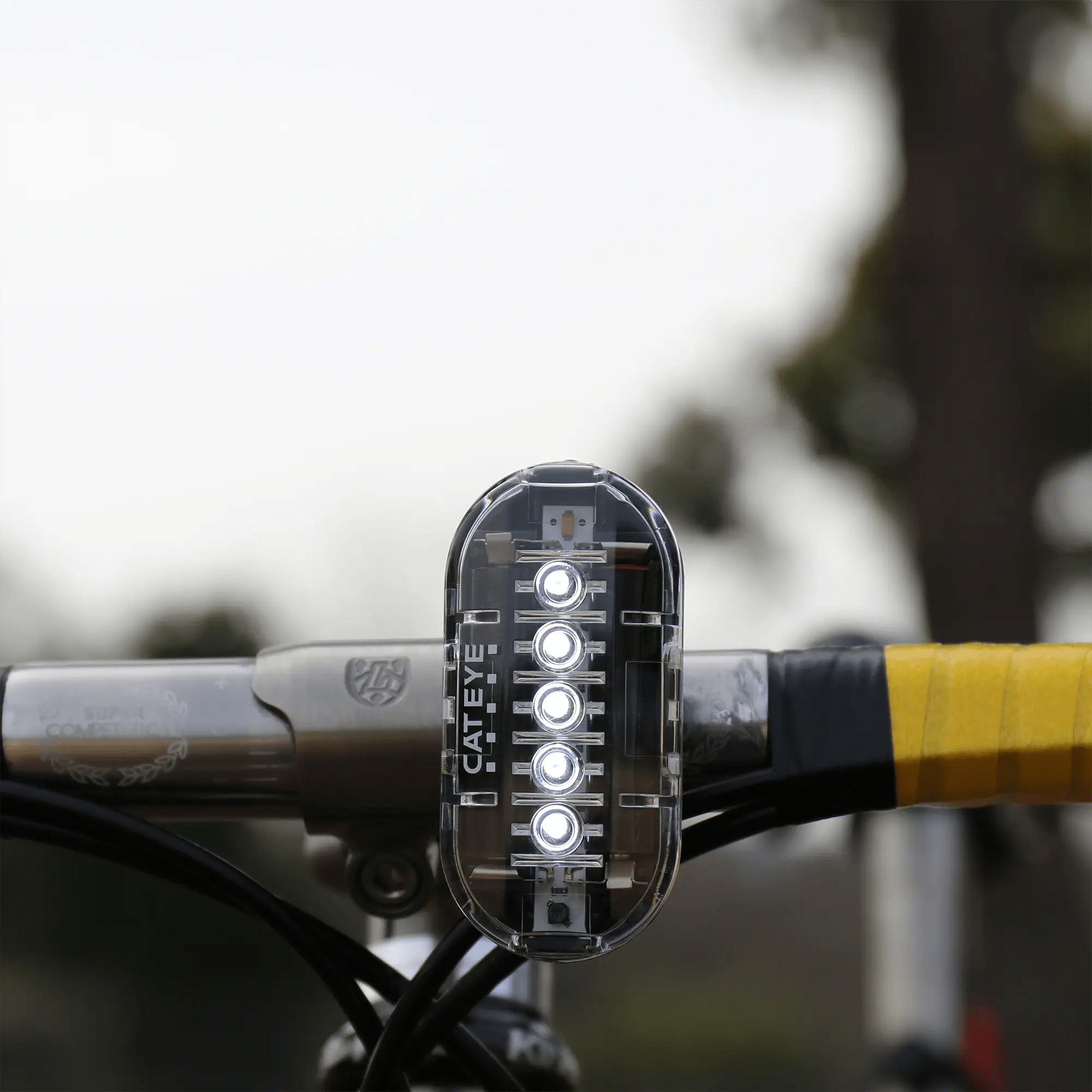 A close-up of the CATEYE OMNI 5 bicycle light, featuring OptiCube™ lens technology and five white LEDs, is mounted on yellow handlebar tape for enhanced brightness and 360° visibility against a blurred outdoor background.