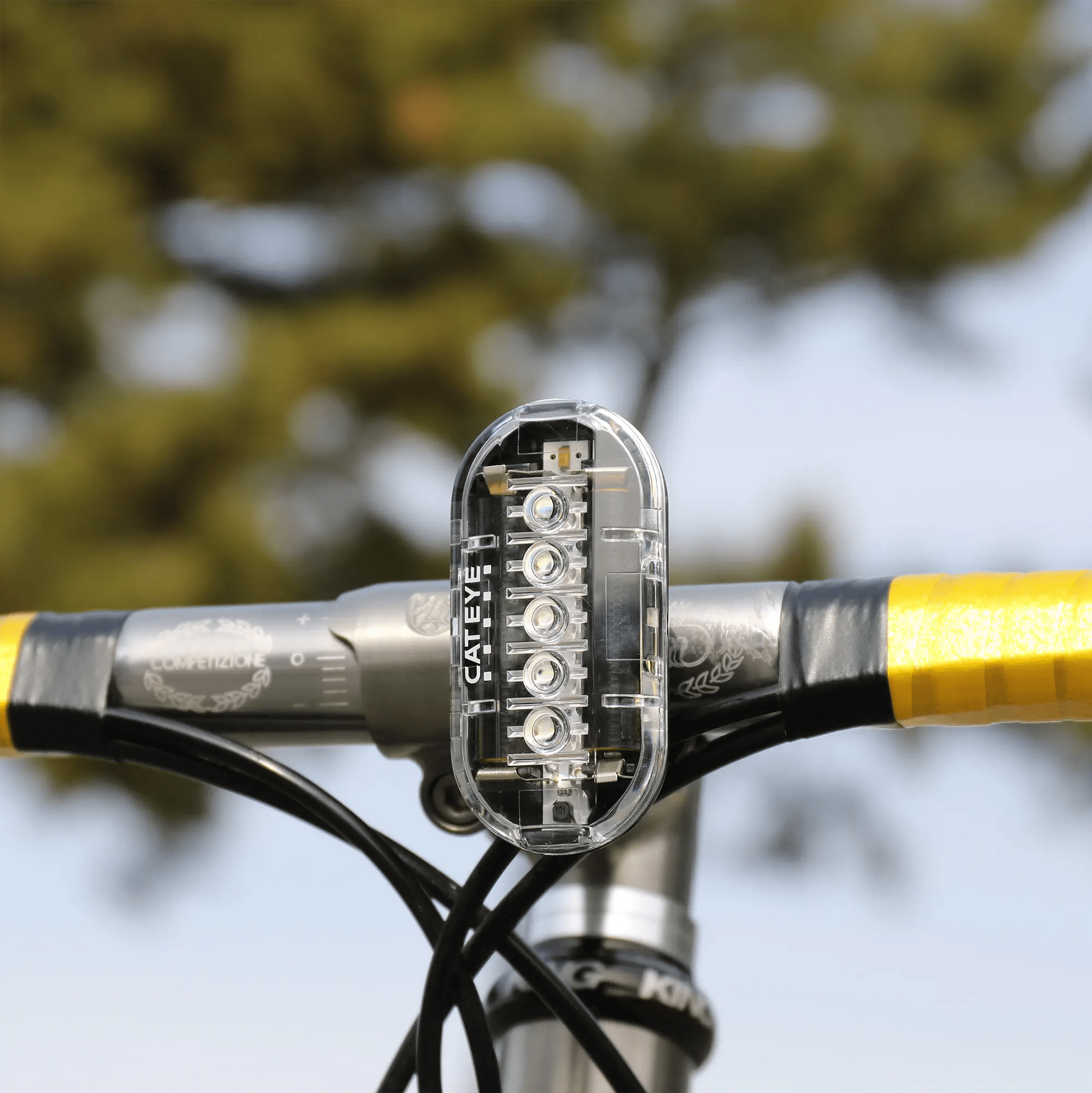 A close-up of a transparent CATEYE OMNI 5 rear light with OptiCube™ lens technology, mounted on handlebars wrapped in yellow tape, offers 360° visibility against a blurred backdrop of green trees and blue sky.