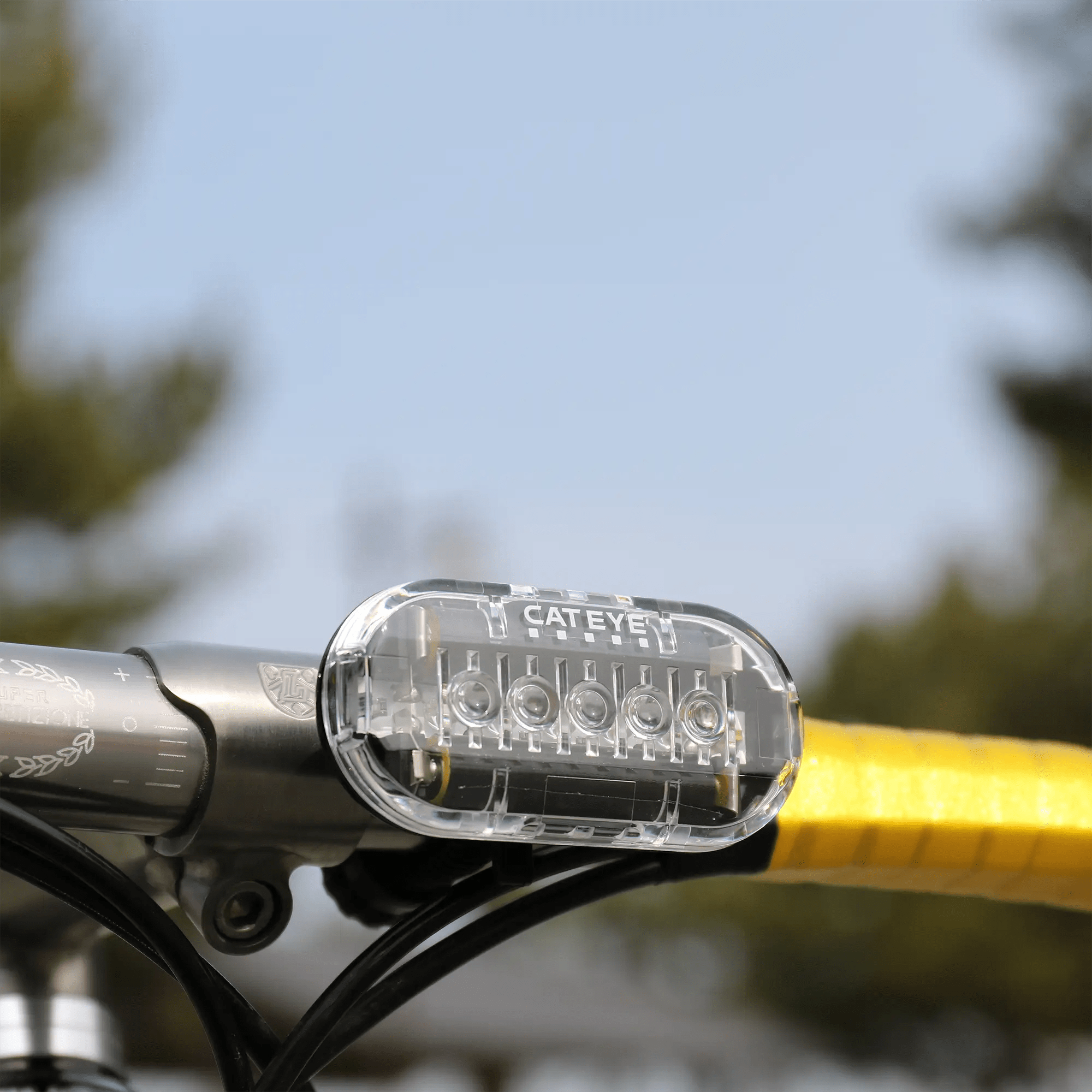 A close-up of a CATEYE OMNI 5 bicycle headlight with OptiCube™ lens technology mounted on yellow handlebar tape, delivering enhanced brightness against a blurred outdoor background.