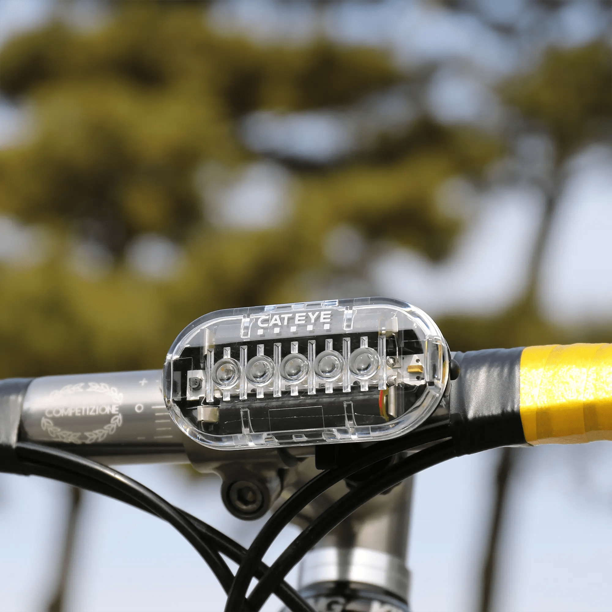 Close-up of a CATEYE OMNI 5 bicycle light with OptiCube™ lens technology mounted on yellow handlebar tape, set against a blurred background of trees.