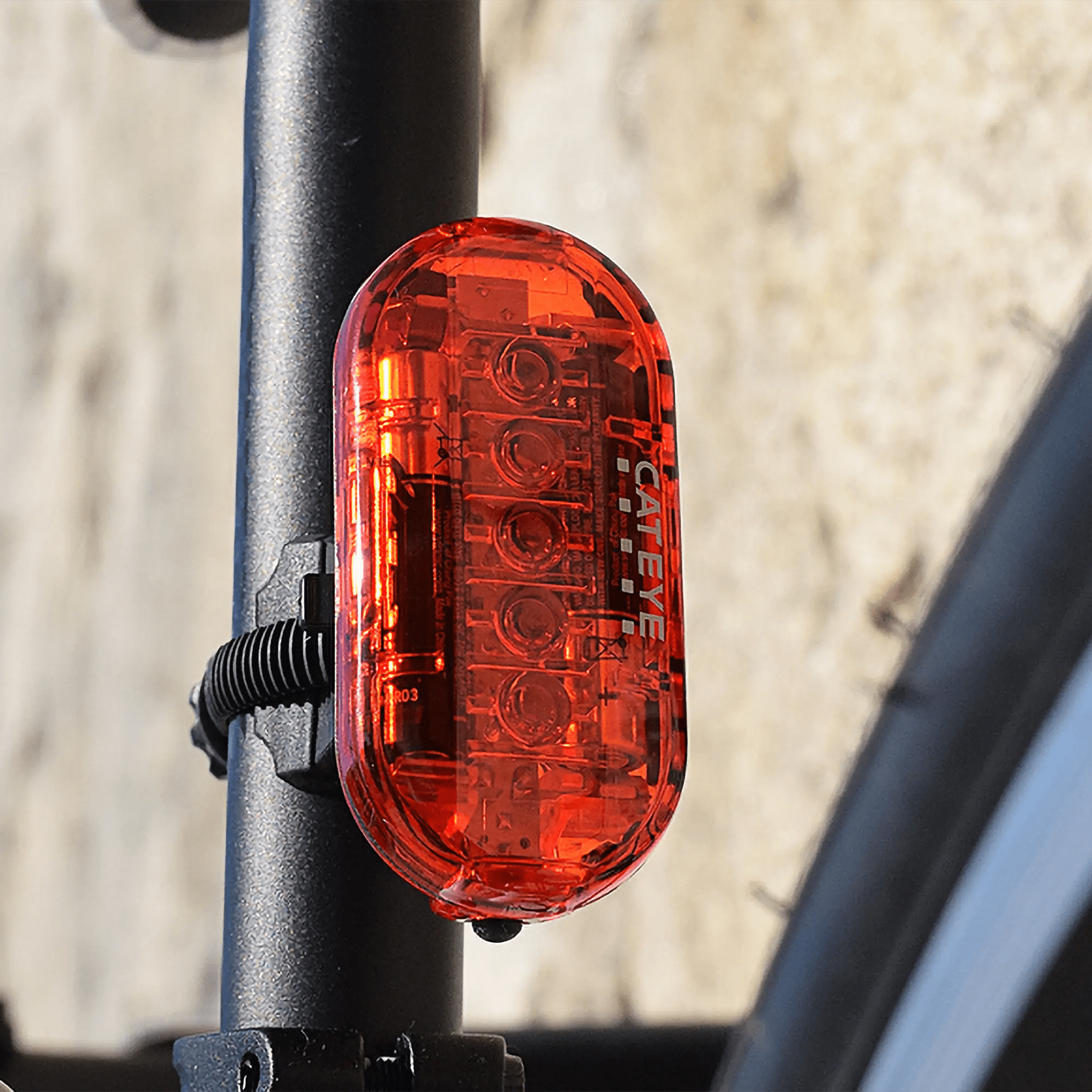 A CATEYE OMNI 5 red rear light with OptiCube lens mounts on the bikes seat post, providing 360° visibility. The rear wheel is partially visible against a blurred outdoor backdrop.