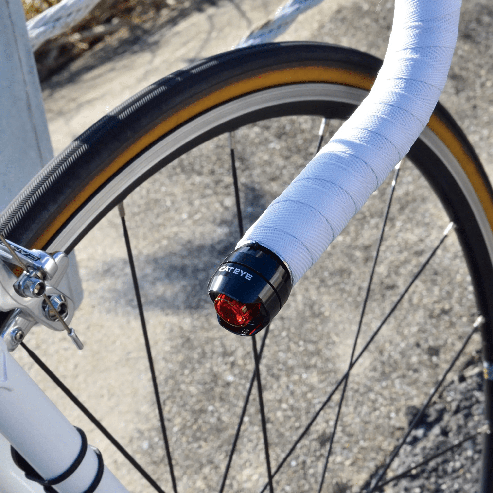 Close-up of a bicycle’s front wheel and white-wrapped handlebars, featuring the CATEYE ORB Bar End safety light for enhanced side visibility. The red rear light offers multiple modes. The bike is parked on a paved outdoor surface.
