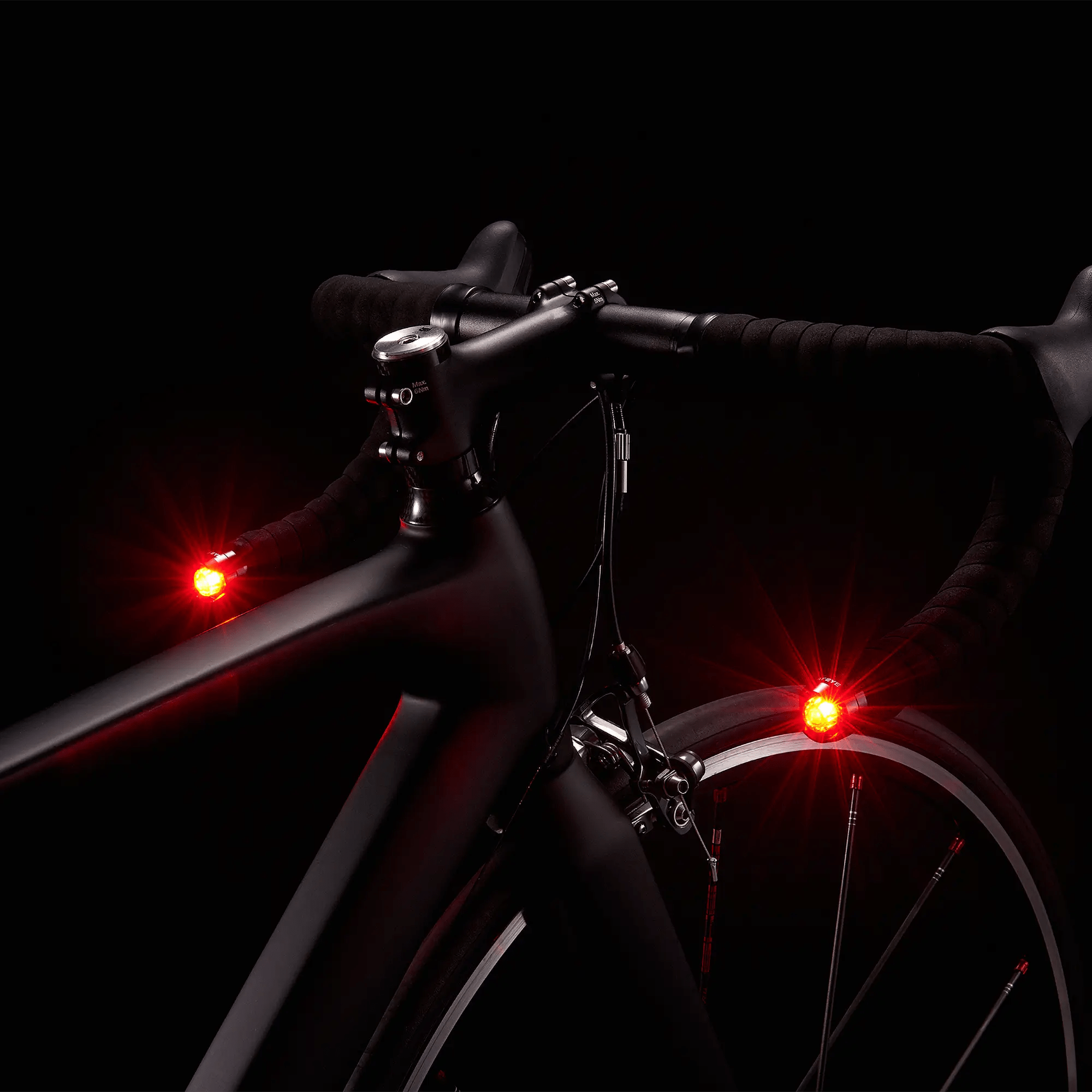 Close-up of a black bicycle with CATEYE ORB Bar End safety lights glowing red on the handlebars and front wheel. Multiple lighting modes enhance side visibility against a dark background.