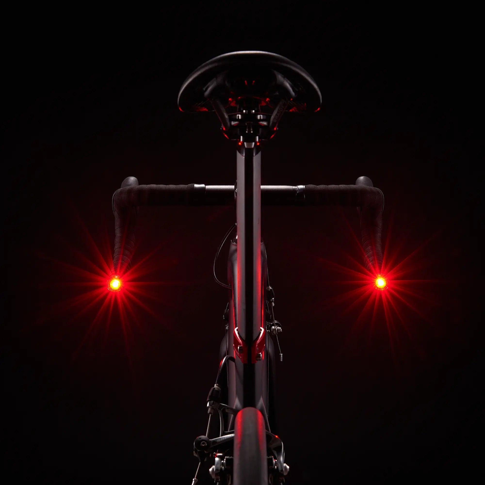 A bicycle is shown from behind in low light with two CATEYE ORB Bar End safety lights on each handlebar end, boosting side visibility as their various lighting modes brighten the darkness.