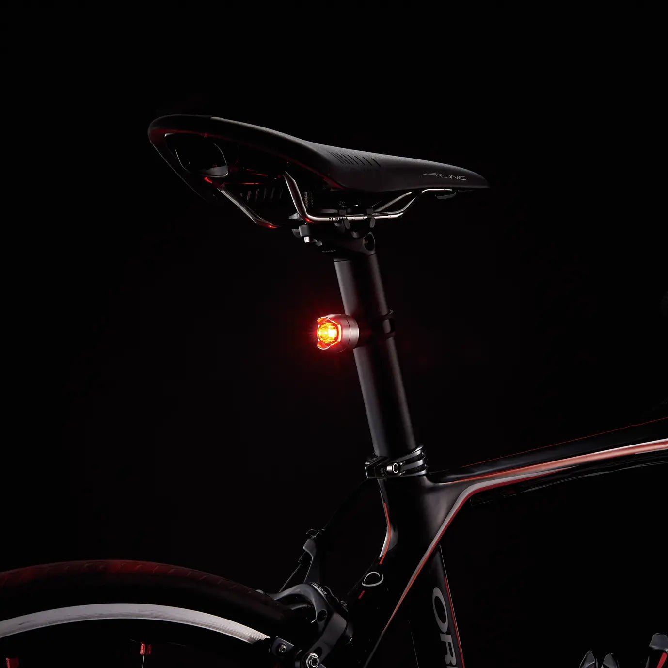 Close-up of the CATEYE ORB bicycle seat and seatpost against a black background, featuring a small red safety light illuminated below the saddle to showcase its essential lighting modes for enhanced visibility.