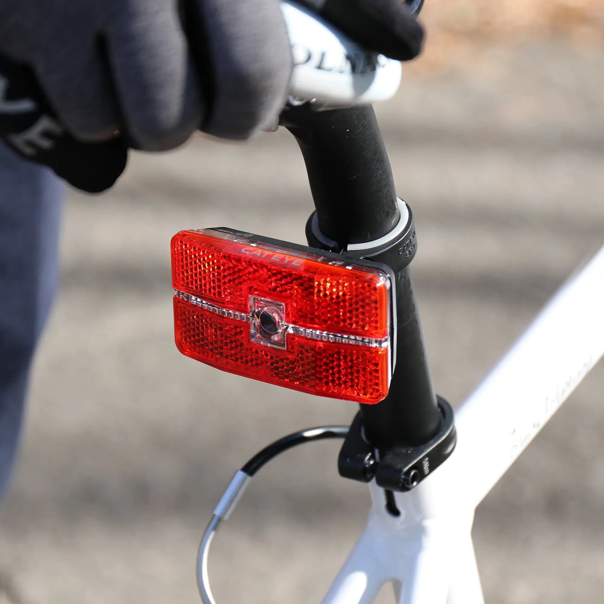 A person wearing gloves grips the handlebars of a white bicycle equipped with the CATEYE Reflex Auto LED tail light, featuring an OptiCube™ lens and red rear reflector on the seat post.