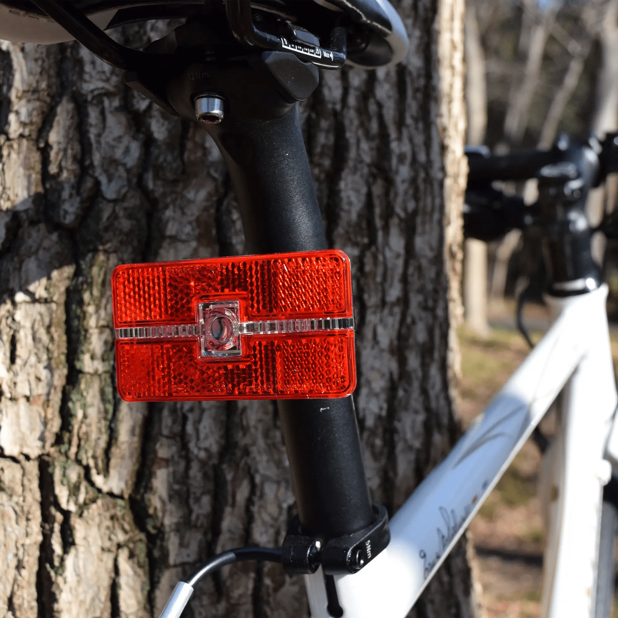 A CATEYE Reflex Auto red reflector or LED tail light is attached to the seat post of a white bicycle parked beside a tree with textured bark outdoors.