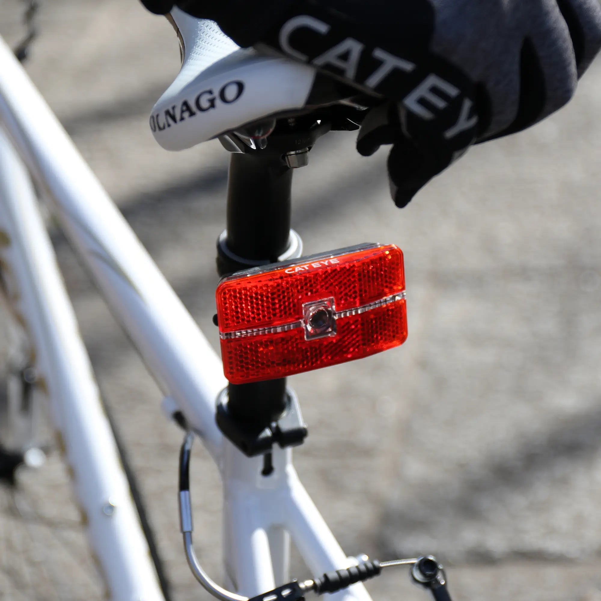 A gloved hand adjusts a CATEYE Reflex Auto LED tail light with an OptiCube™ lens, mounted under the seat of a white Colnago bike. The glove shows CATEY, and the background is blurred pavement.