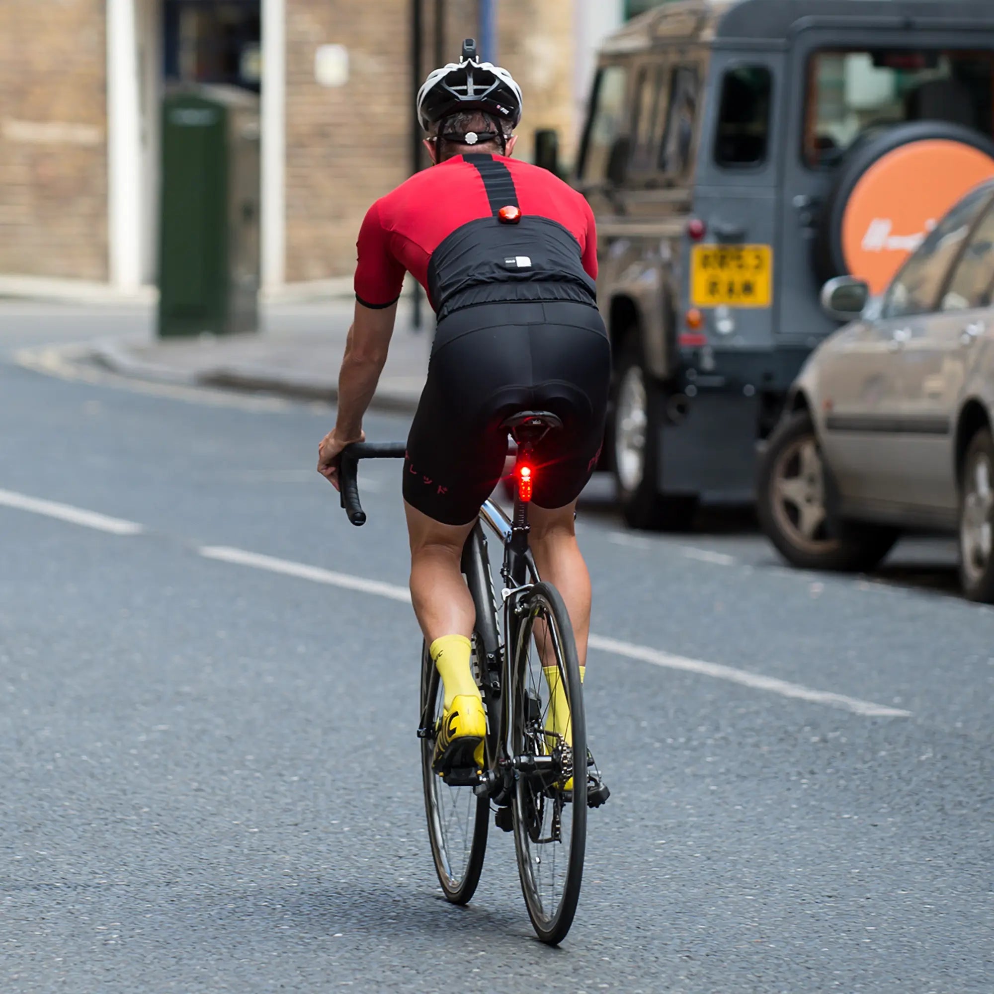 A cyclist in red and black with yellow socks rides a road bike on a city street. The CATEYE SYNC Kinetic rear light uses CatEyeSYNC wireless tech to boost 360-degree visibility near parked cars.