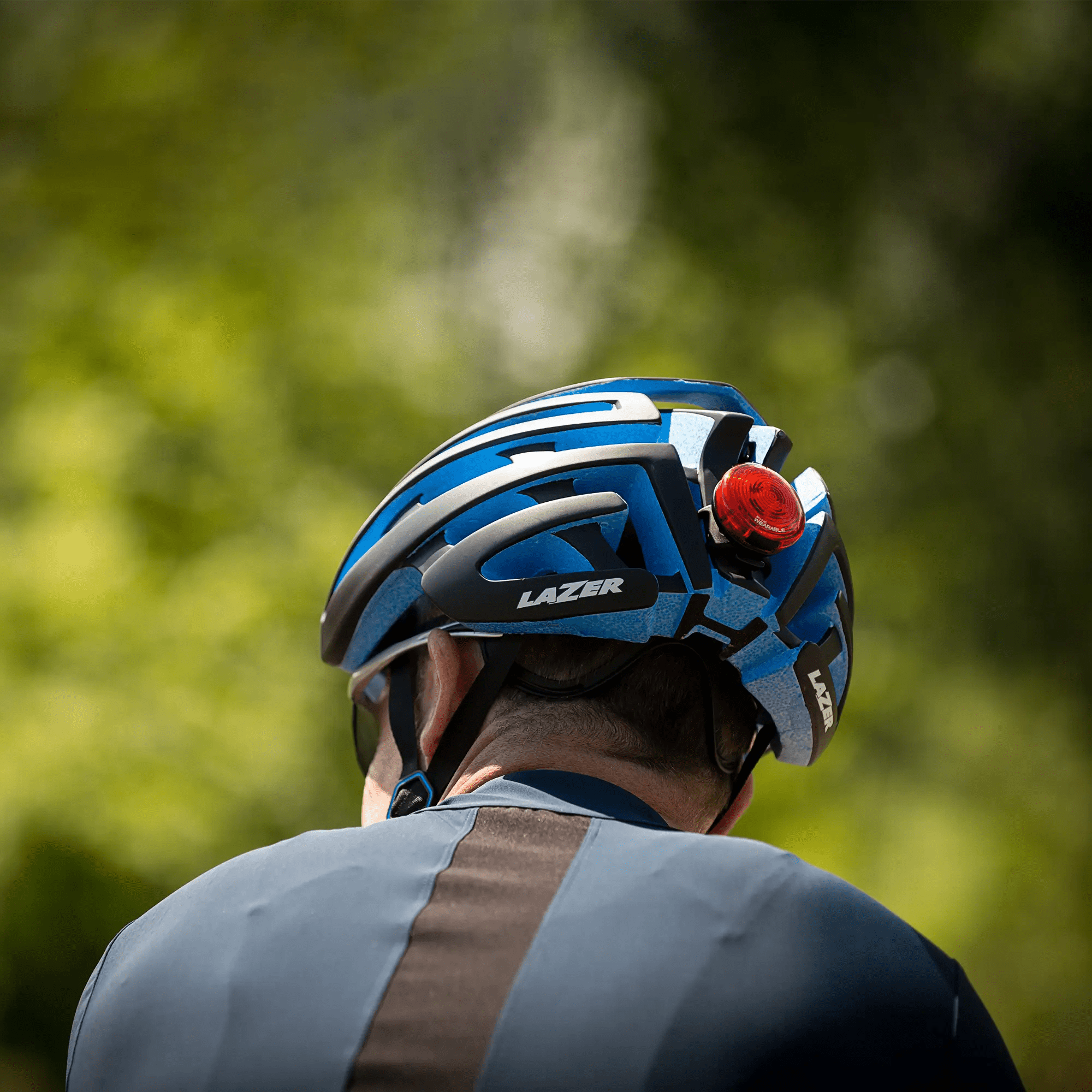 A person is seen from behind wearing a blue and black Lazer bike helmet with a red CATEYE SYNC Wearable rear light for enhanced 360-degree visibility, set against a blurred green background.
