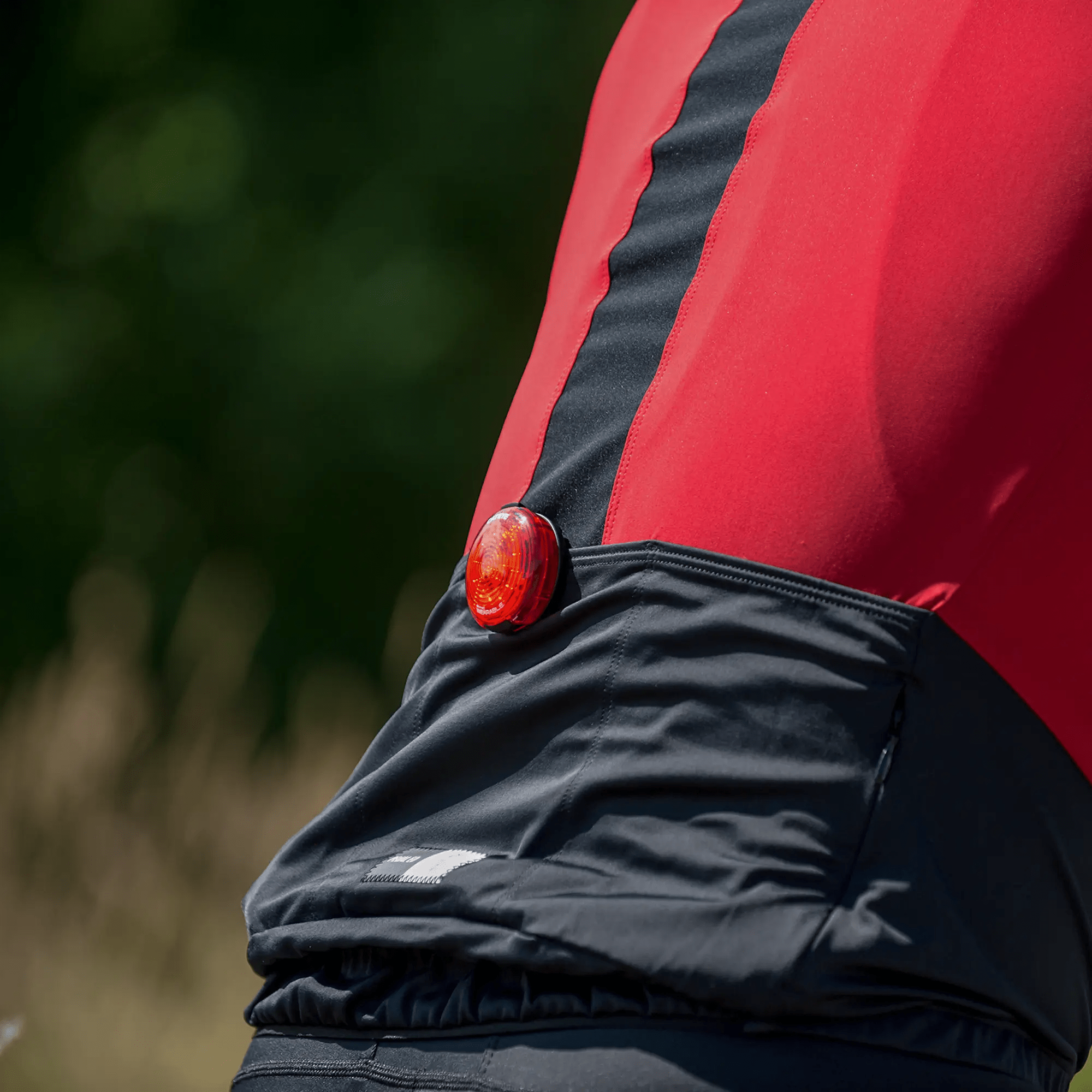 A person wearing a red and black cycling jersey has the CATEYE SYNC Wearable rear bike light clipped to their back pocket for 360-degree visibility. The blurred background features greenery.