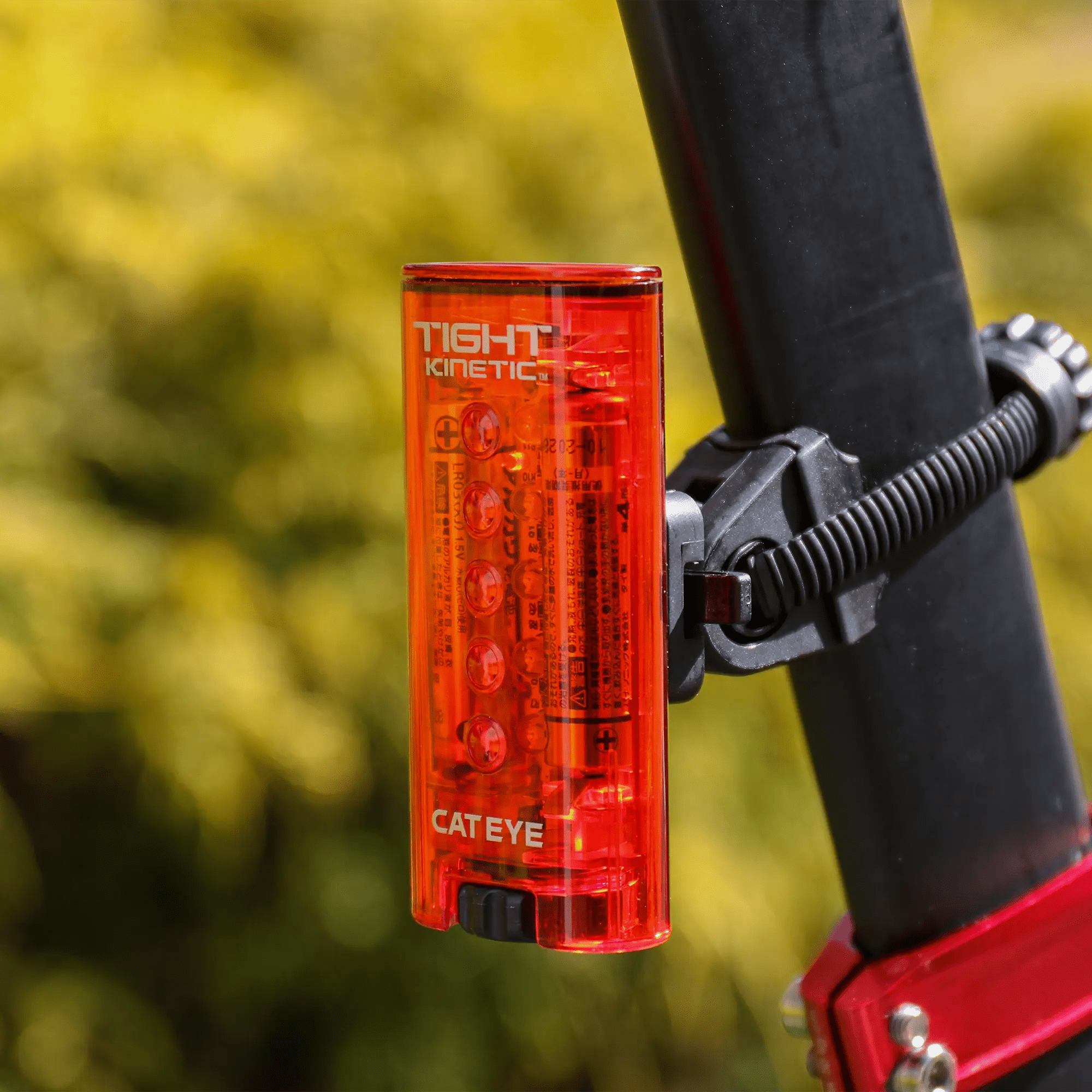 A CATEYE Tight Kinetic red rear light with an acceleration sensor is attached to a black seat post with a clamp, set against a blurred green outdoor background.