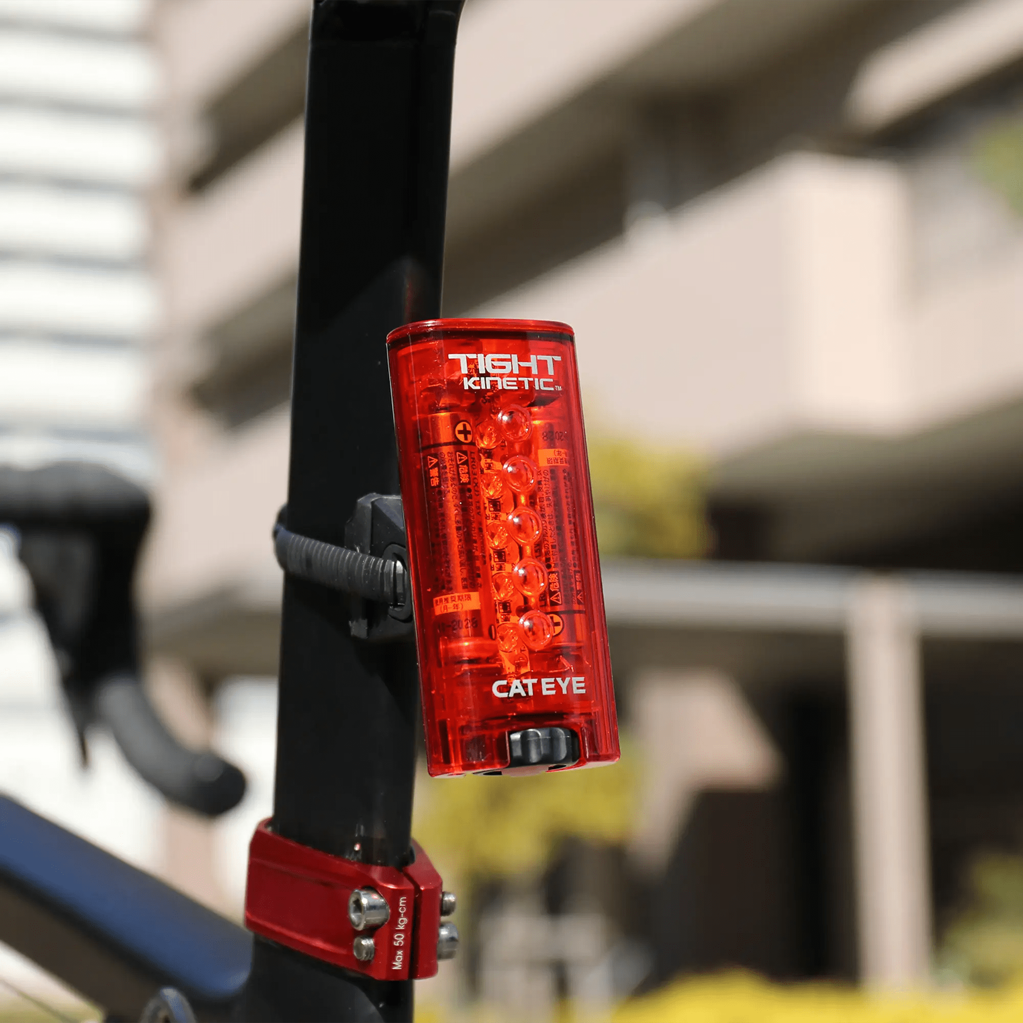 A CATEYE Tight Kinetic rear light with IPX7 waterproof rating is mounted on a bicycle seat post, with blurred buildings and greenery in the background.