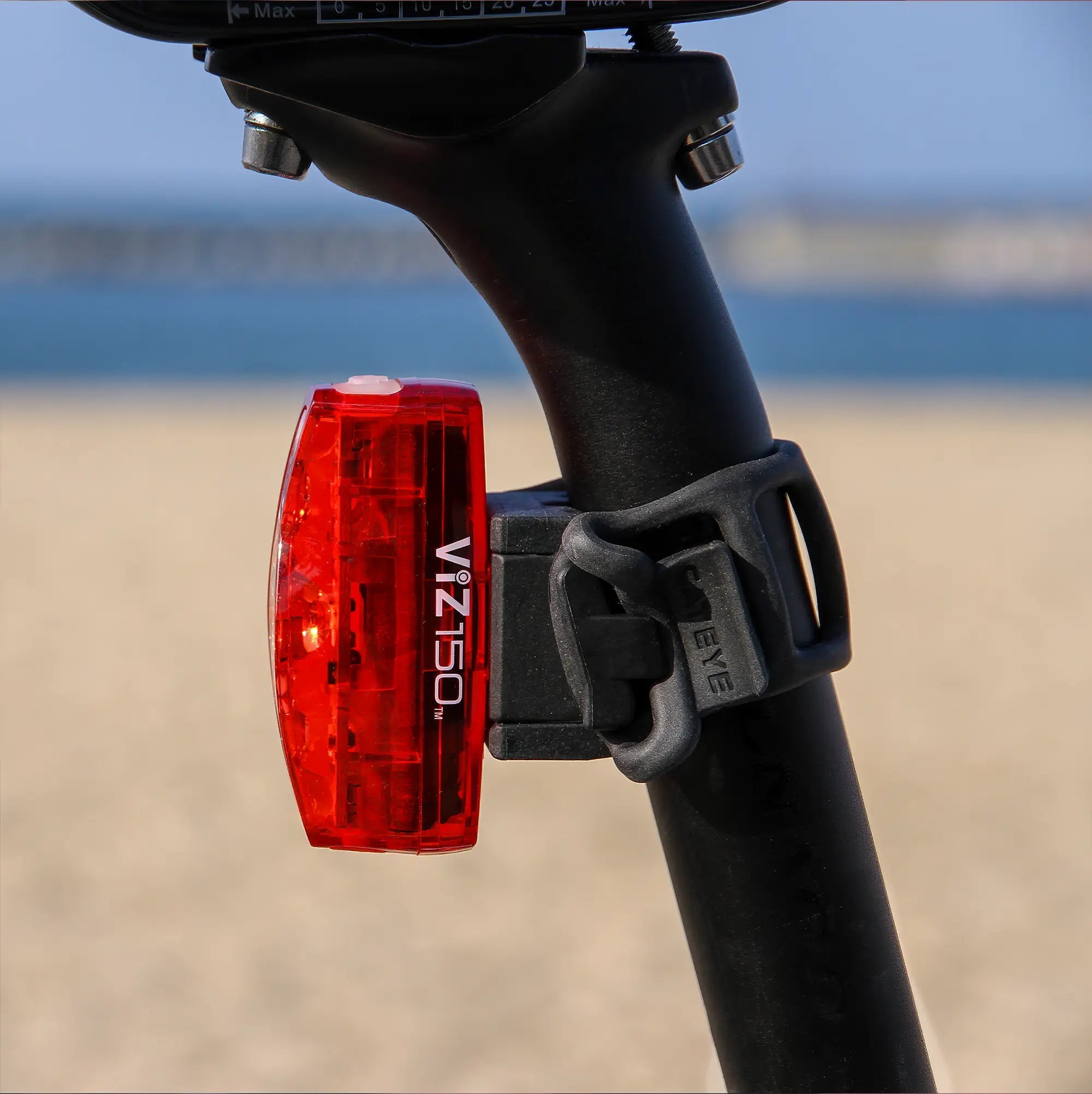 A CATEYE ViZ150 (USB-C) red LED bike light is mounted on a black seatpost with a rubber strap. The blurred backdrop of sand and blue water suggests a beach setting. OptiCube™ lens technology ensures enhanced visibility.