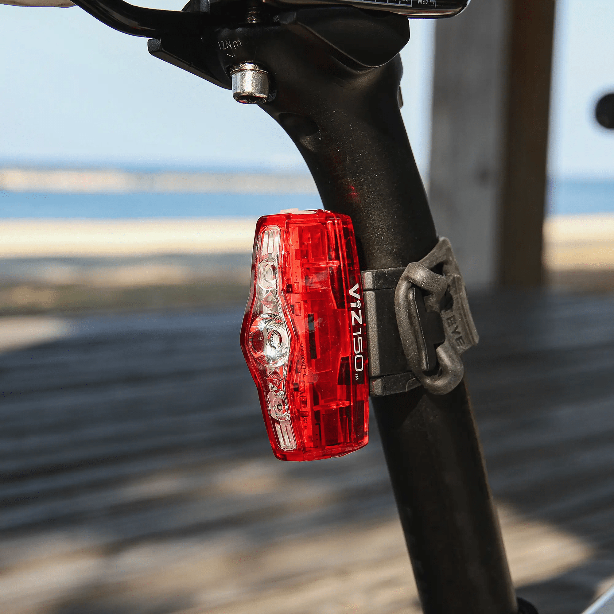 A CATEYE ViZ150 (USB-C) red LED bike light with OptiCube™ lens technology is mounted on the seat post of a black bike, with a blurred boardwalk and water in the background.
