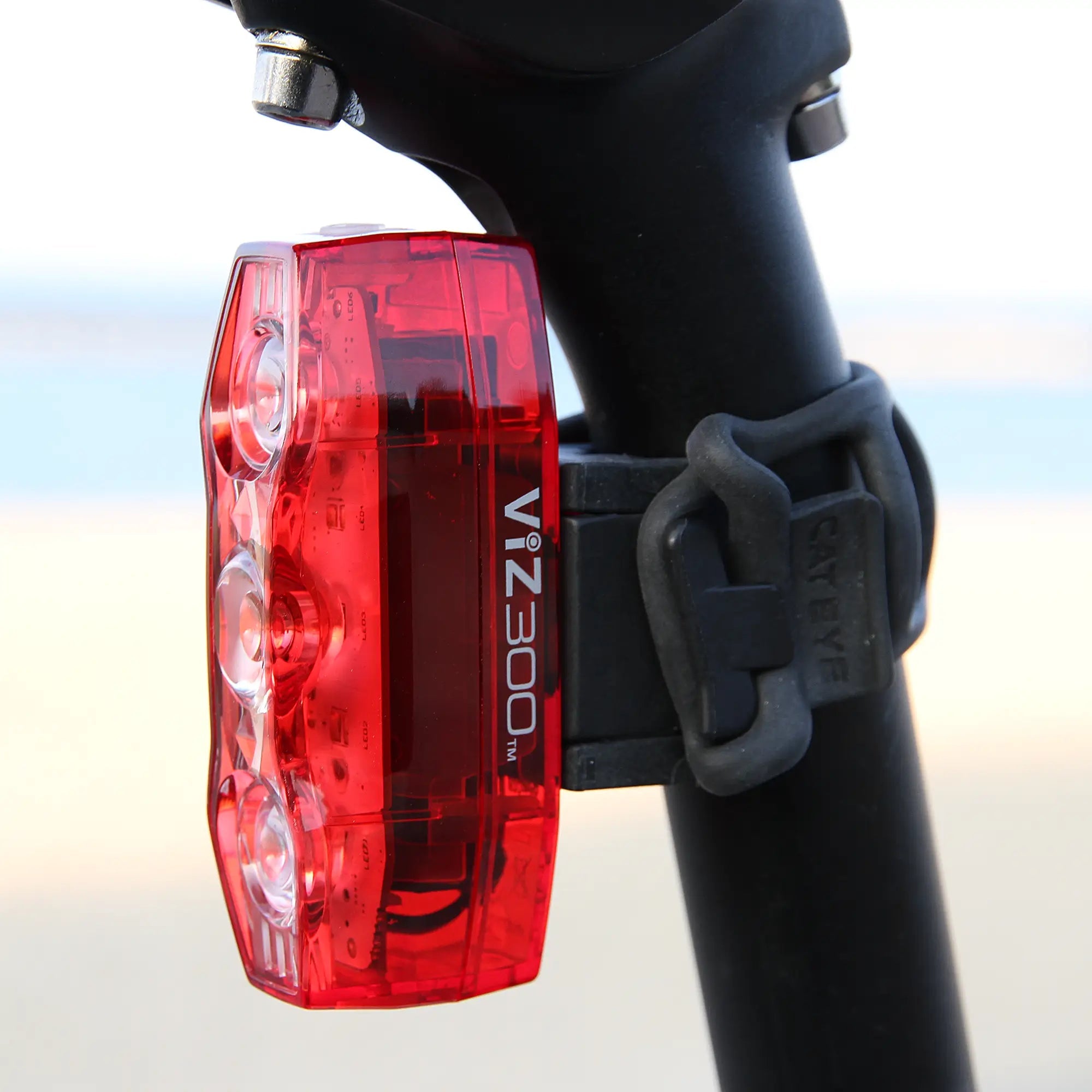 A CATEYE ViZ300 (USB-C) red bike light is mounted to a black seat post with a rubber strap outdoors, background blurred. It offers enhanced visibility with Daytime Hyperflash™ mode.