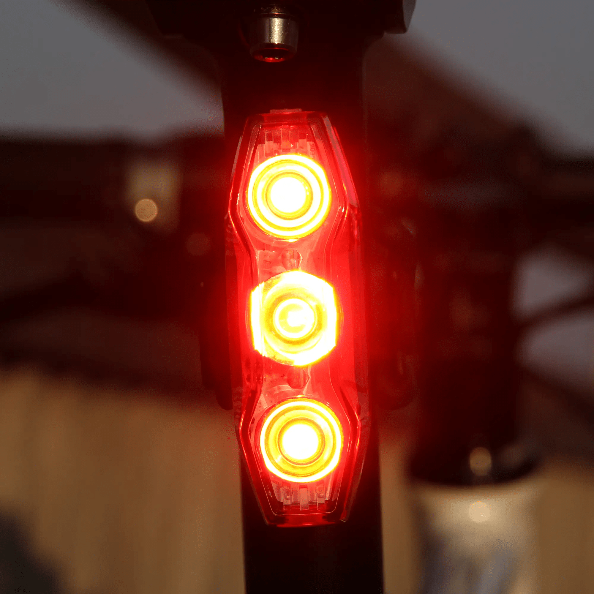 Close-up of the CATEYE ViZ300 (USB-C) bike light with three red LED rings glowing in Daytime Hyperflash™ mode, mounted on a seat post for enhanced visibility in low-light. The background appears softly blurred.