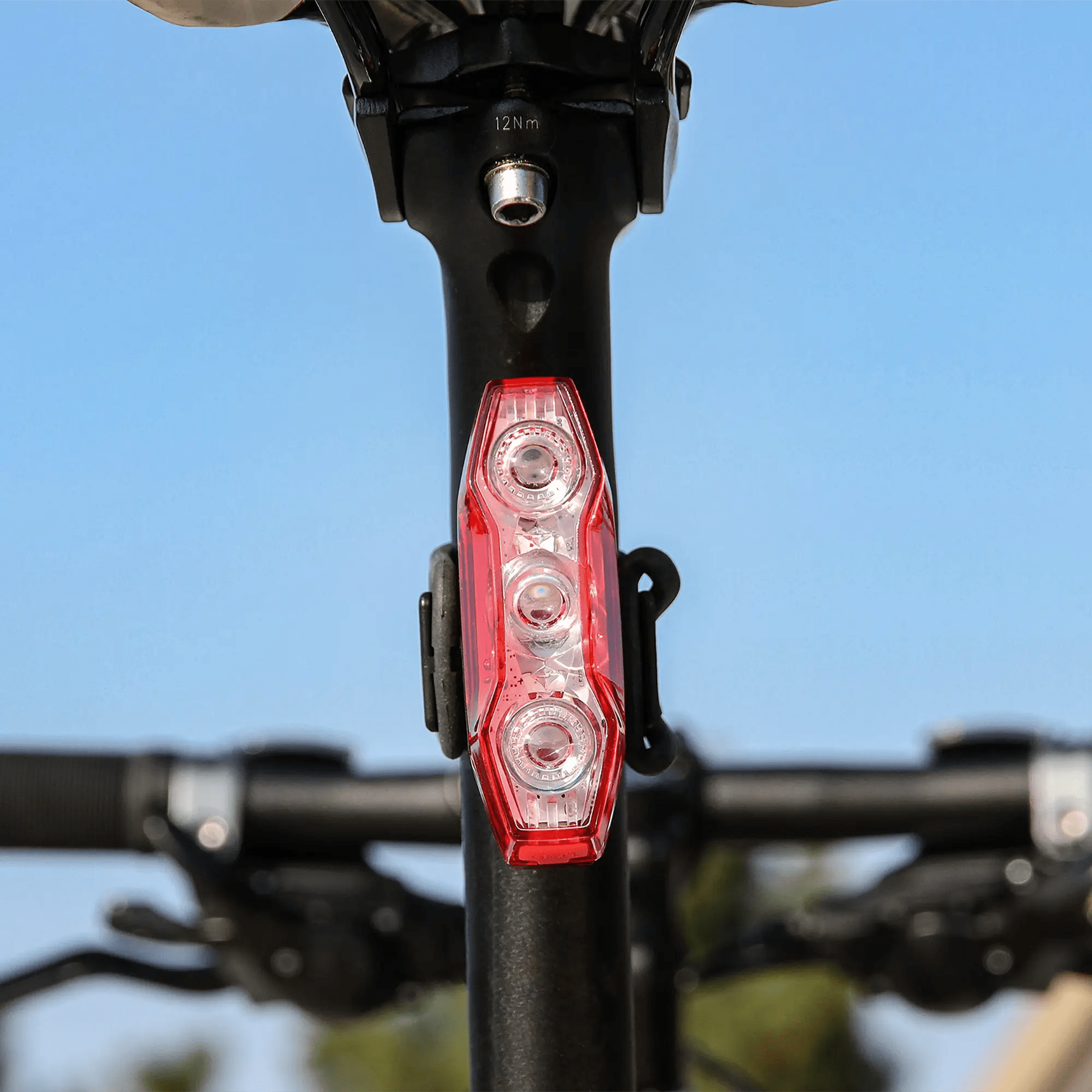 Close-up of the CATEYE ViZ450 (USB-C) rear safety light with three LED bulbs and OptiCube™ lens technology, mounted on a seat post. Blurred handlebars and clear blue sky in the background.