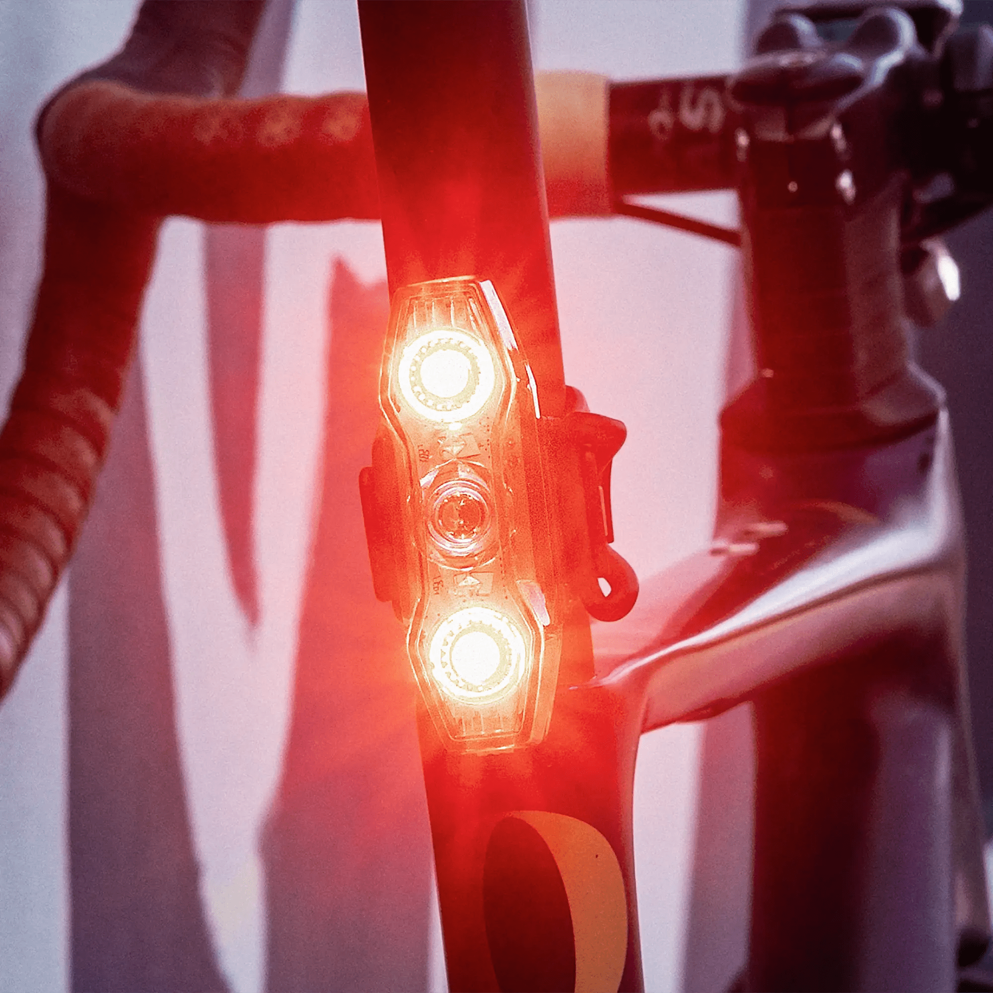 A close-up of the CATEYE ViZ450 (USB-C) rear safety light with OptiCube™ lens tech, glowing brightly with two LEDs on a seat post. The focus is on the light, while handlebars and frame are slightly blurred in the background.