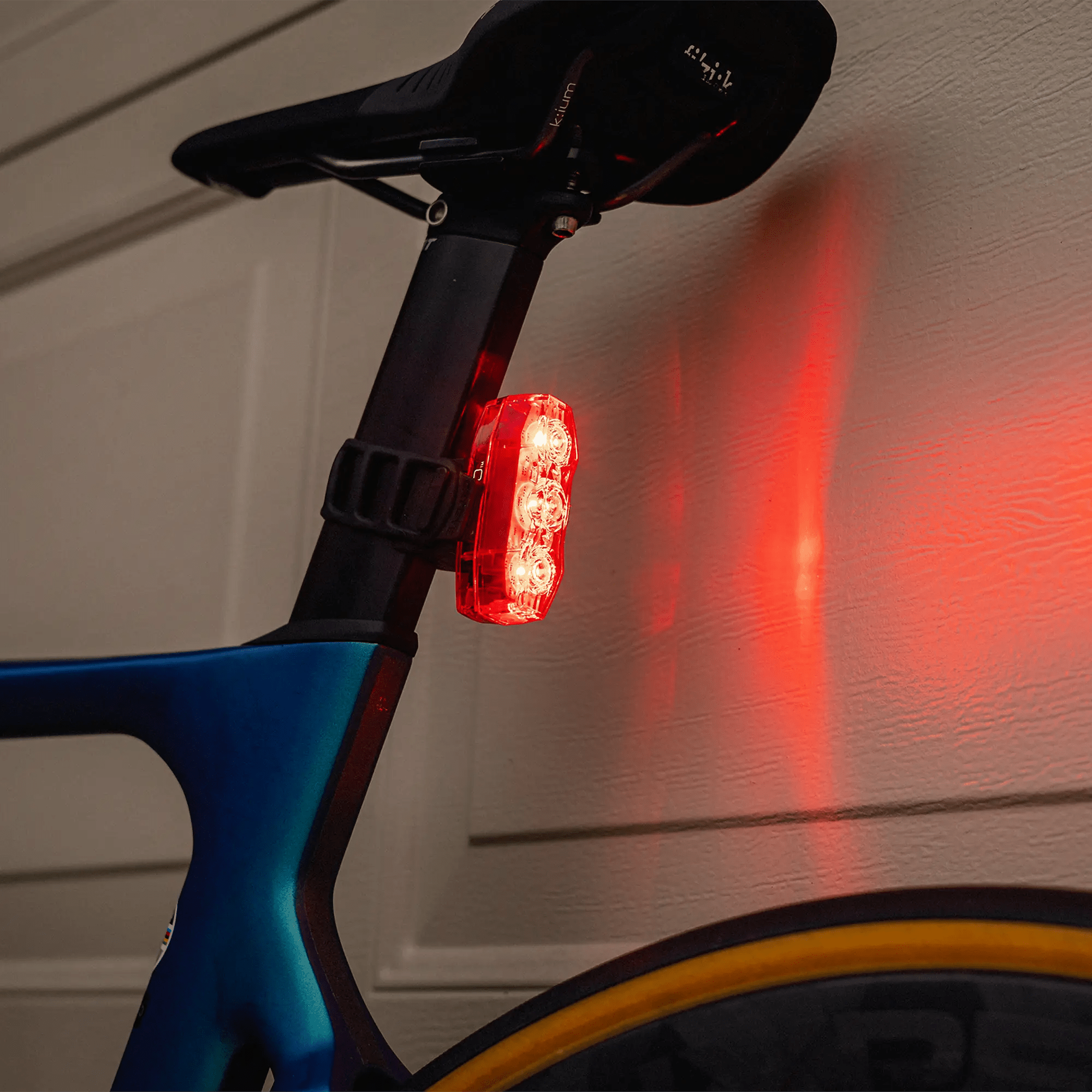 A close-up of CATEYE’s ViZ450 (USB-C) rear safety light with OptiCube™ lens casts a red glow onto a beige garage door. The bike frame is blue, and a yellow-rimmed tire appears at the bottom.
