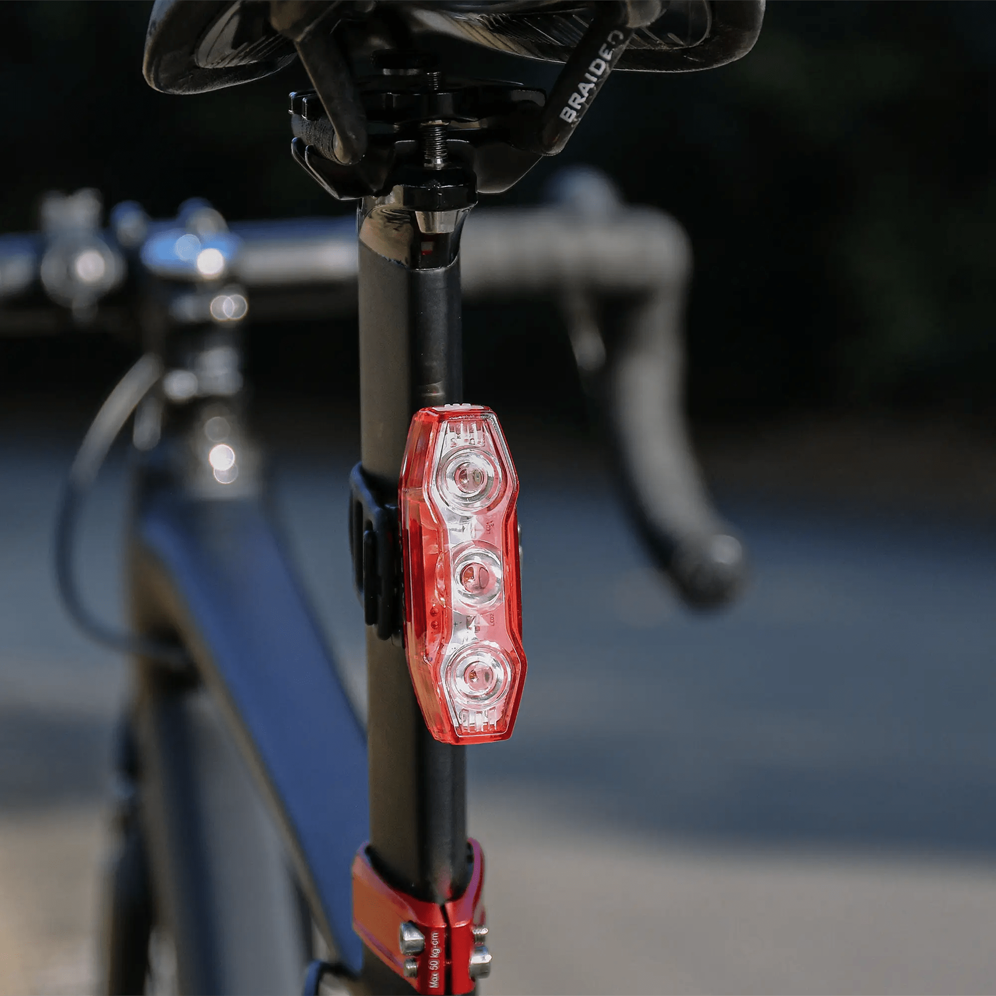 A close-up of the CATEYE ViZ450 (USB-C) red rear safety light with three LEDs and OptiCube™ lens, mounted on a black bicycle’s seat post, with handlebars blurred in the background.