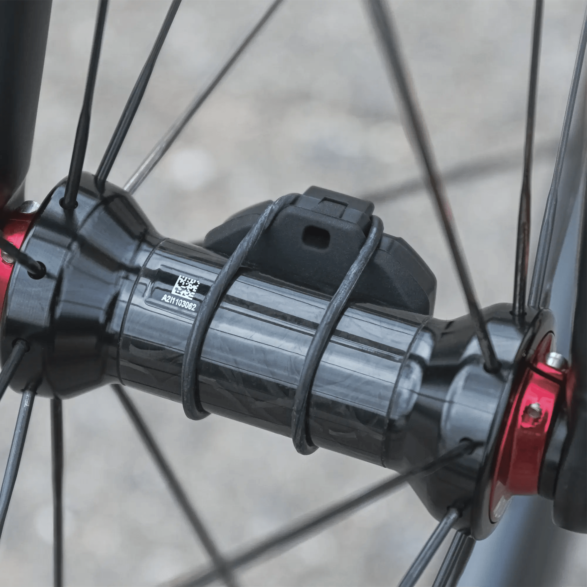 Close-up of a CATEYE SPD-30 Magnetless Speed Sensor with Bluetooth® and ANT+ connectivity, zip-tied to a bicycle wheel among black spokes and red hub ends, against a blurred outdoor background.