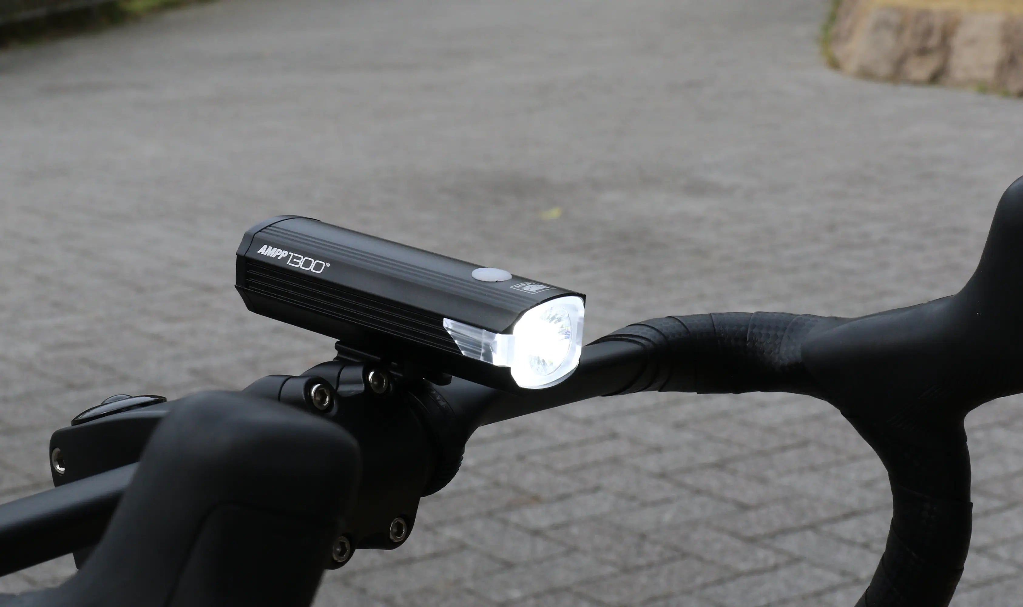 A close-up of a black bicycle handlebar with a bright front bike light turned on, mounted securely, with a paved surface in the background.