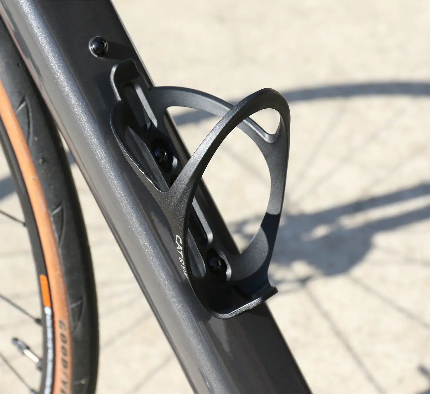 Close-up of a CATEYE BC-400 Bottle Cage mounted on a black bicycle frame; part of the tire and shadow appear on the light-colored ground in the background.