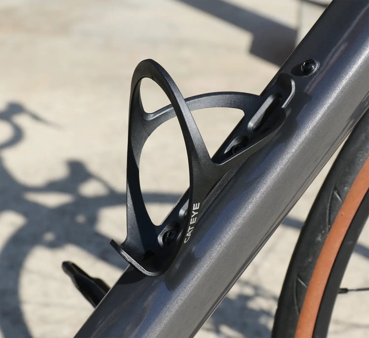 Close-up of a black CATEYE BC-400 Bottle Cage mounted on a bicycle frame, with a blurred background and the bike’s shadow visible on the ground.
